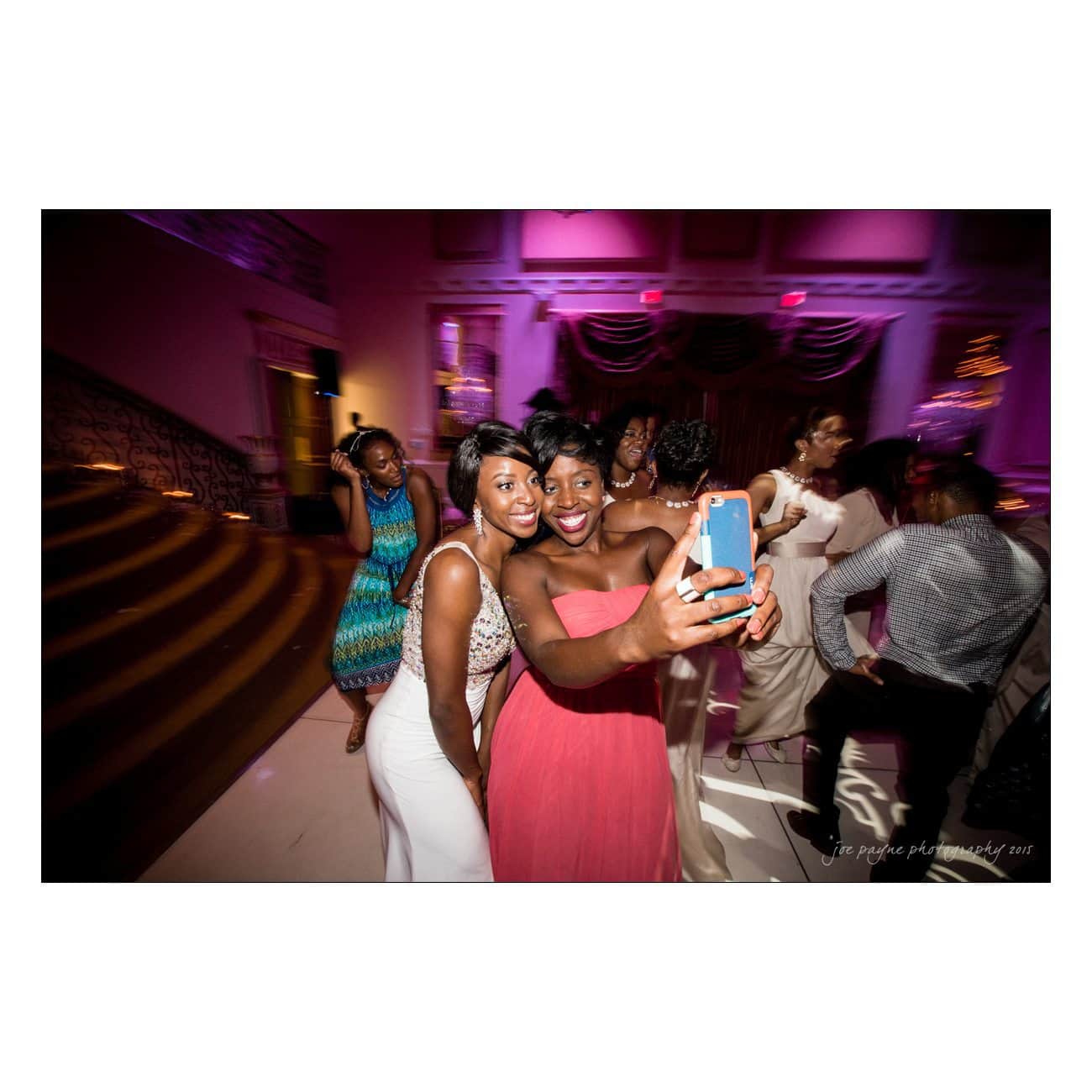 Two people wearing elegant dresses take a selfie at a lively event. The scene is filled with people, vibrant lights, and a grand staircase in the background, creating a festive atmosphere.