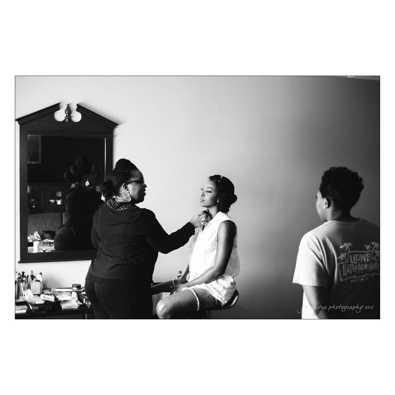 Two women are applying makeup on someone seated in front of a mirror. The setting appears to be a dressing room. A person is standing nearby, watching. The atmosphere is relaxed and focused on preparation.