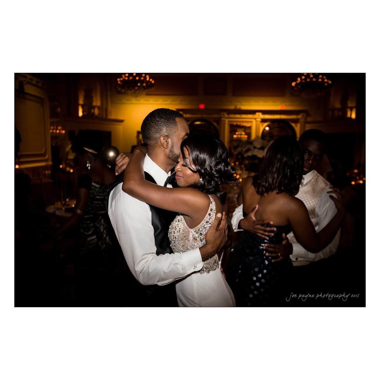 A man and woman are embracing while dancing in a dimly lit ballroom. The room has elegant decor with chandeliers. Other couples are visible in the background.
