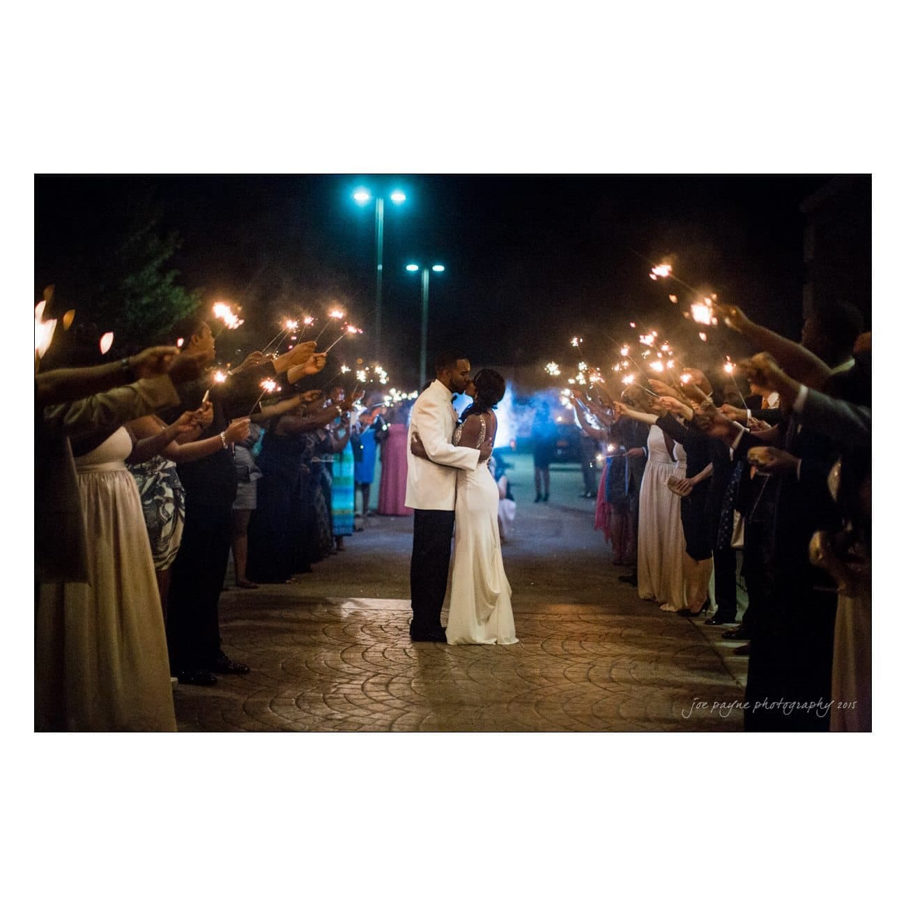 A couple dressed elegantly shares a kiss under a night sky, surrounded by people holding sparklers. The scene is romantic, with the glowing lights creating a magical atmosphere.