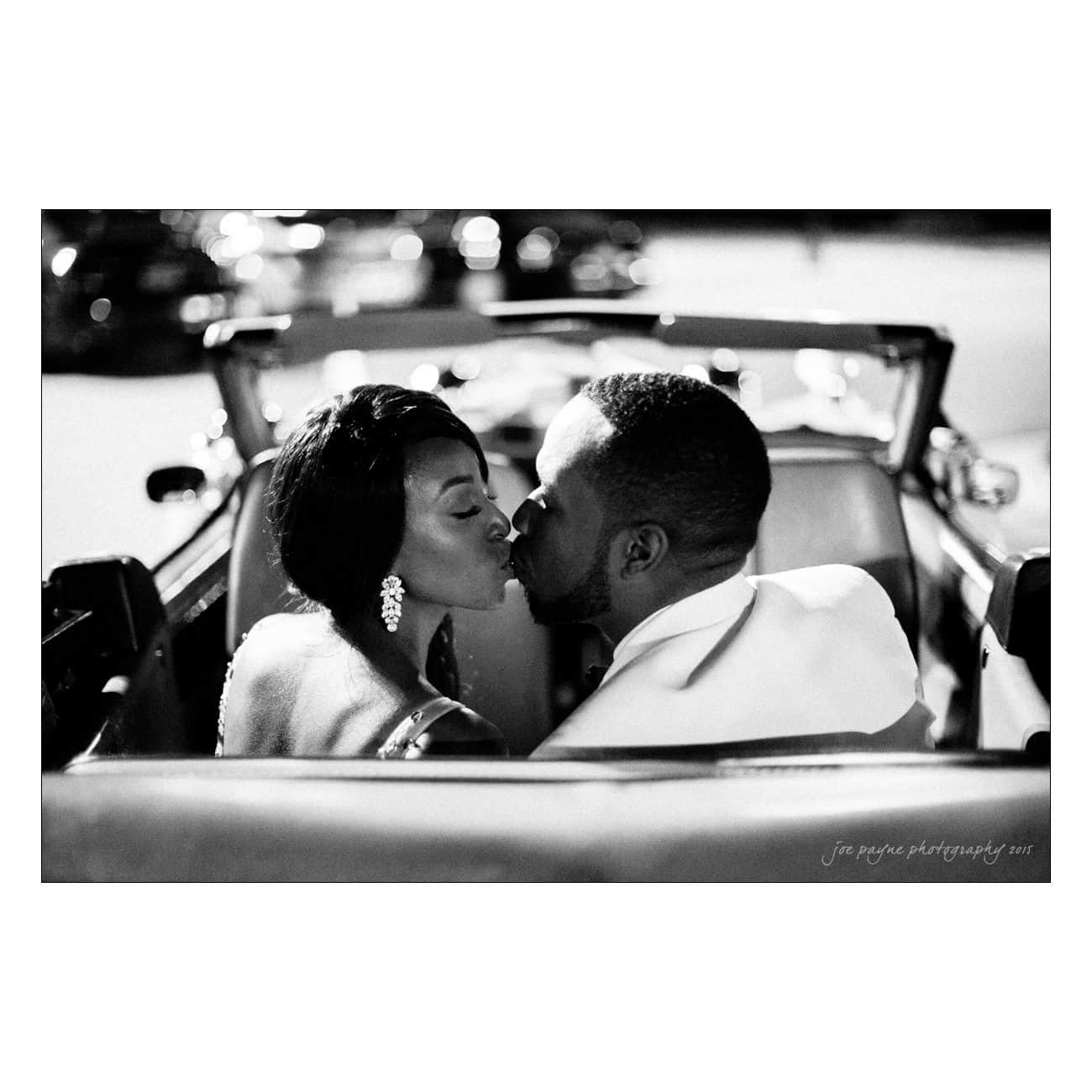 A couple seated in a convertible car shares a kiss, captured in black and white. They are dressed elegantly, with the woman wearing earrings and the man in a suit. The focus is on their intimate moment at night.