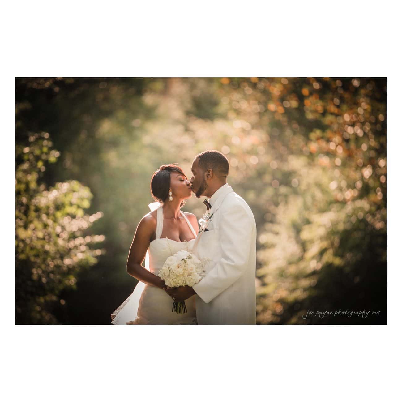 A bride and groom dressed in white share a kiss outdoors at their grand marquise wedding, surrounded by soft sunlight and greenery, with the bride holding a bouquet of white flowers.