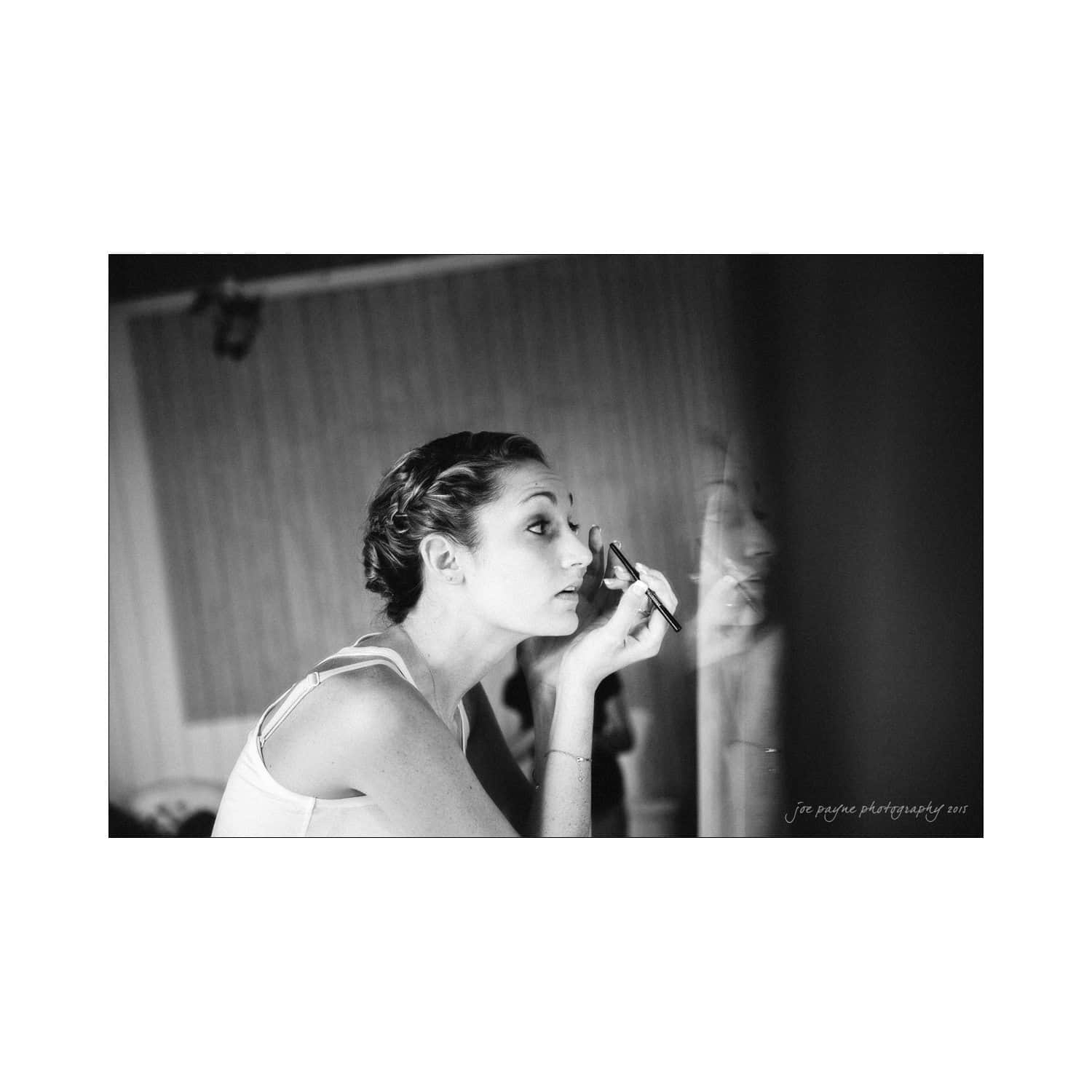 chapel hill wedding photography - nicole & wayne Black and white photo of a woman applying makeup in front of a mirror. She has braided hair and is focusing on her reflection while holding a makeup brush. The background is softly blurred.
