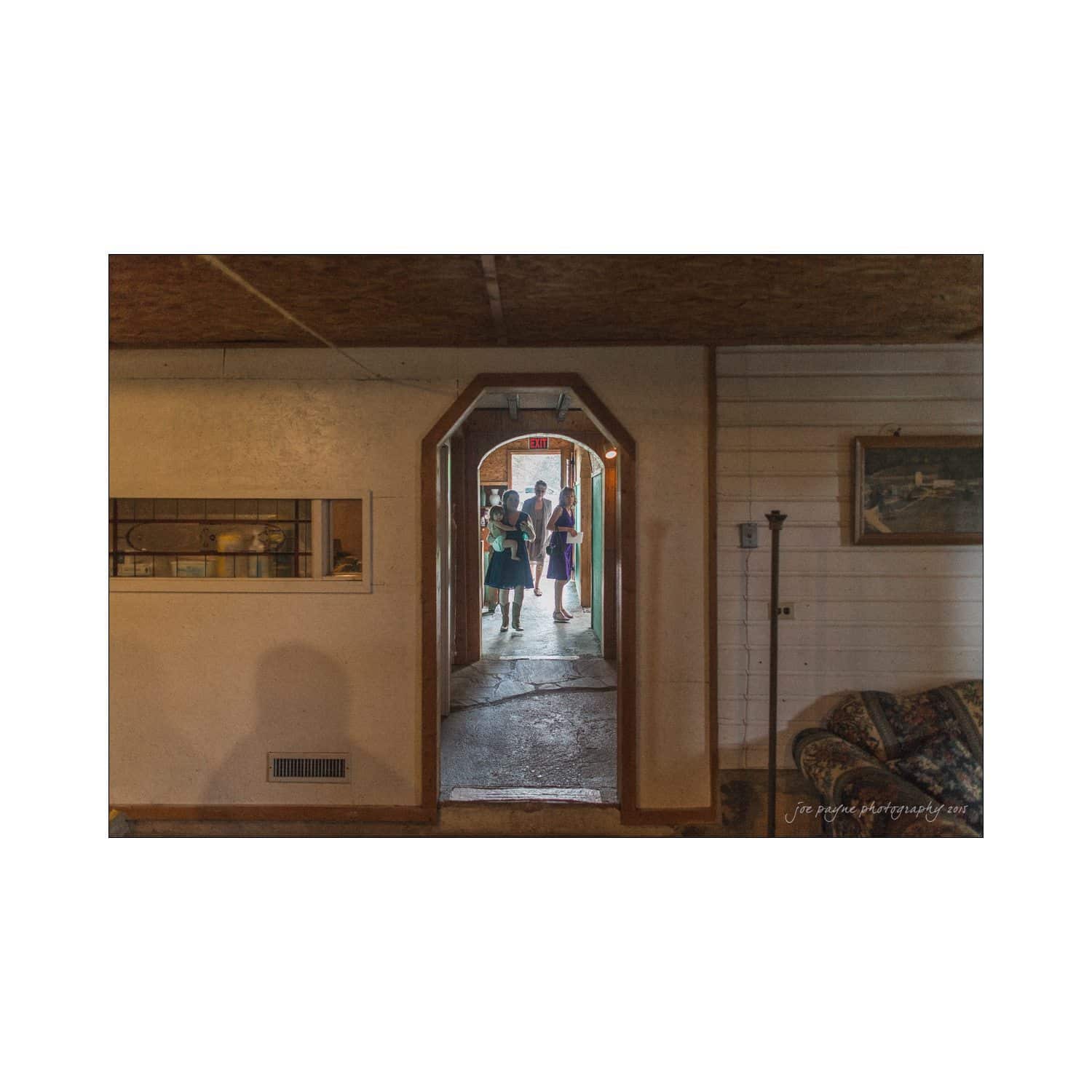 chapel hill wedding photography - nicole & wayne A view through an arched doorway shows three people entering a house. The interior features a cozy room with a sofa and framed artwork on the walls. The atmosphere is warm and inviting, with natural light streaming in from outside.