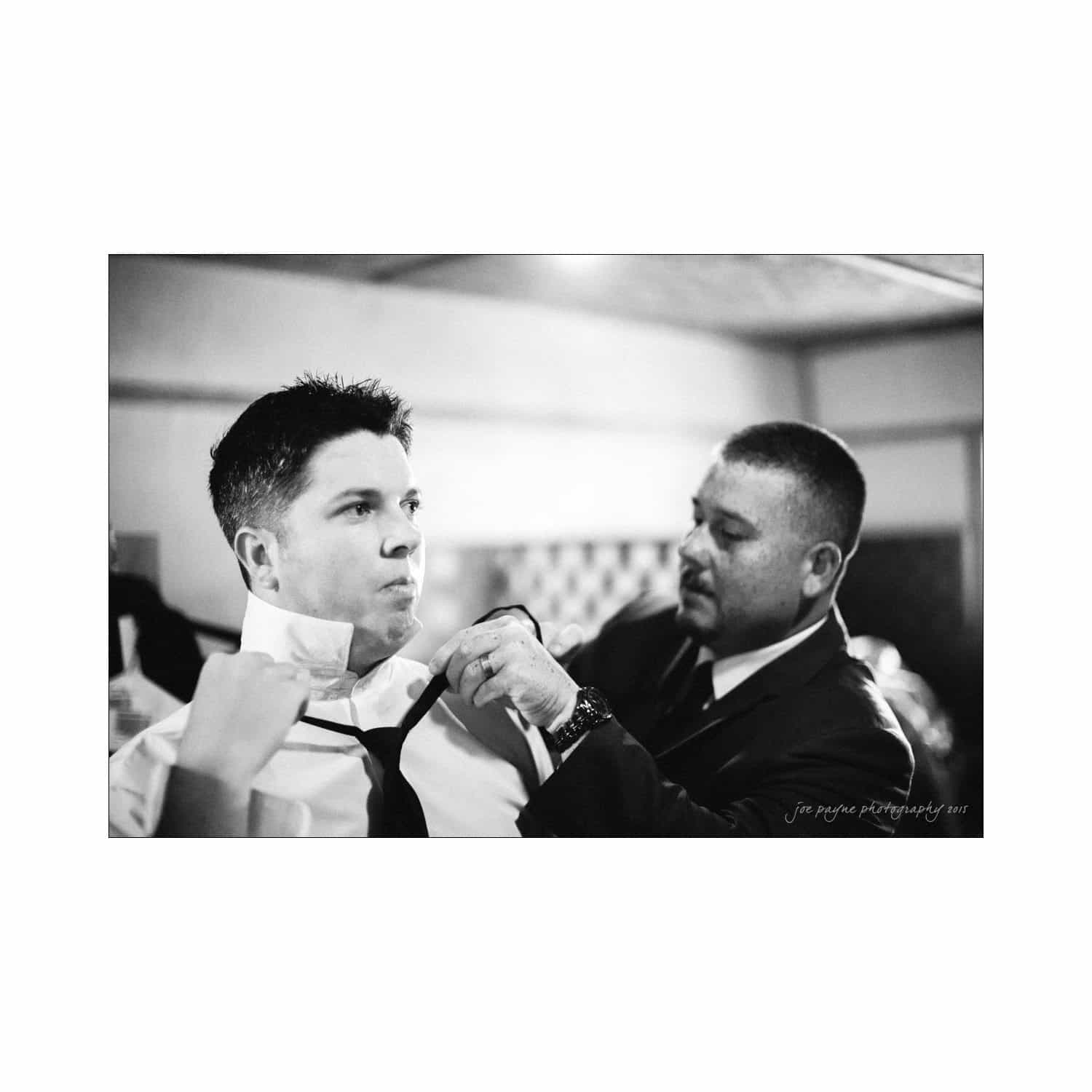 chapel hill wedding photography - nicole & wayne A man helps another man adjust his tie in preparation for an event. They are in a softly lit indoor setting, and both appear focused and dressed in suits. Black and white photograph.