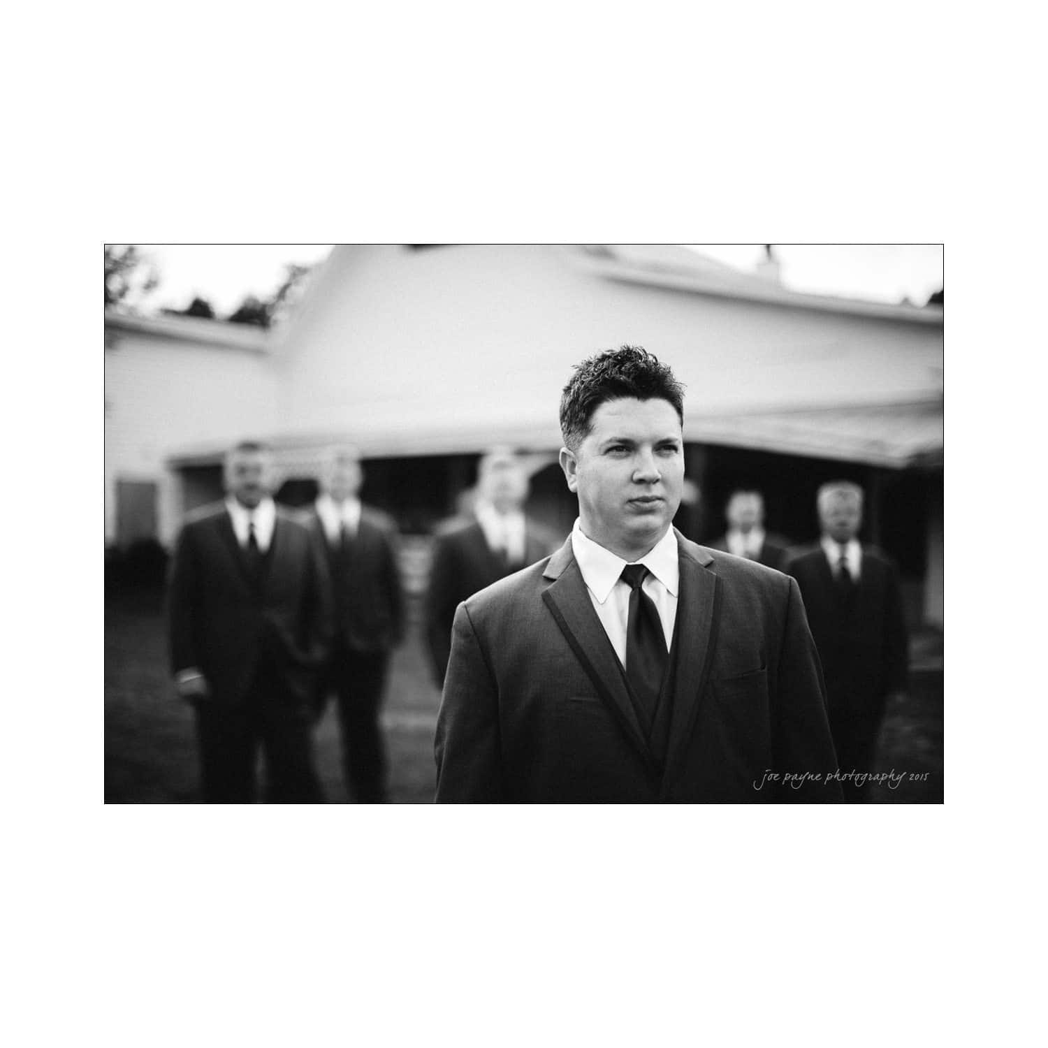 chapel hill wedding photography - nicole & wayne A black and white photo of a groom in a suit standing outside, surrounded by groomsmen in blurred background. They are all wearing suits and ties. A barn-like building is visible in the background.