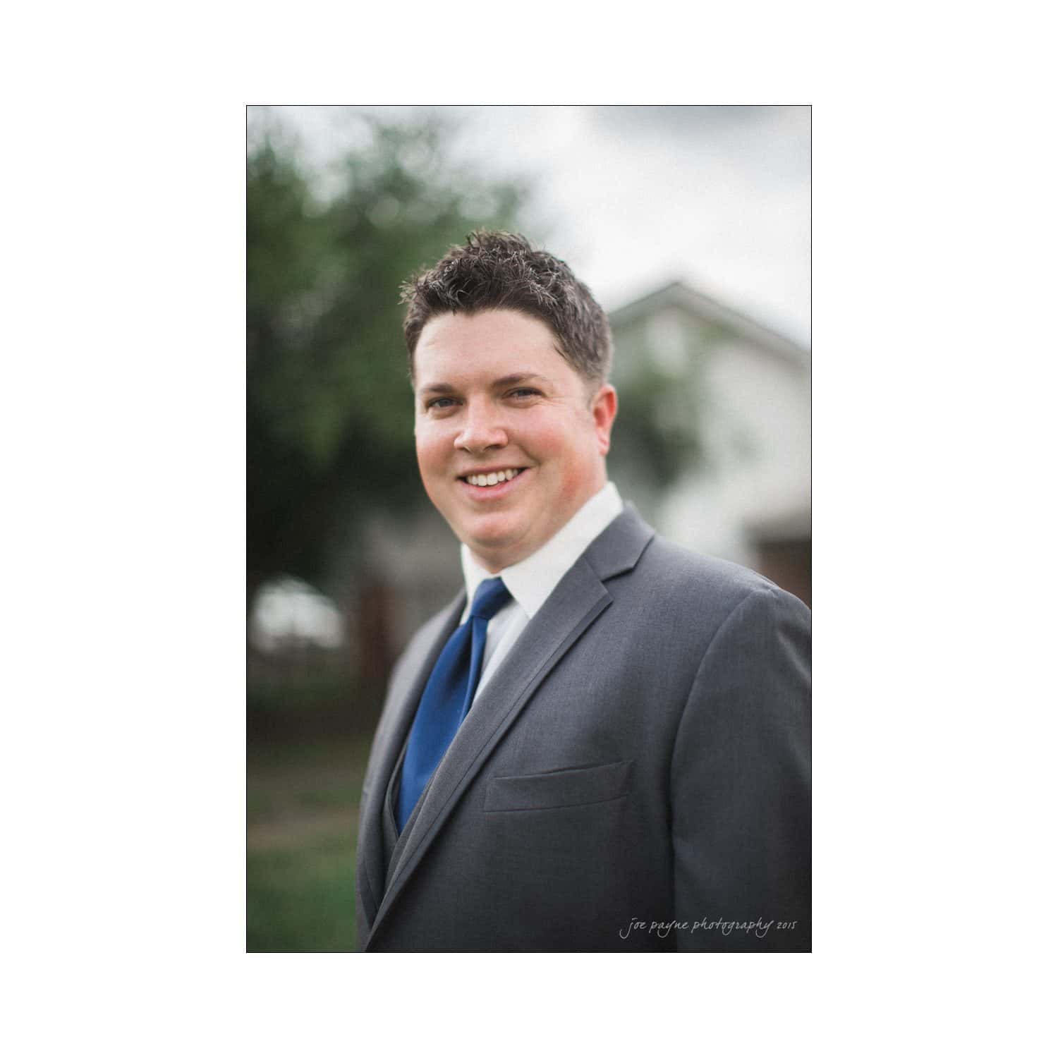 chapel hill wedding photography - nicole & wayne A man in a gray suit and blue tie is smiling outdoors. The background is blurred, showing green foliage and a white building.