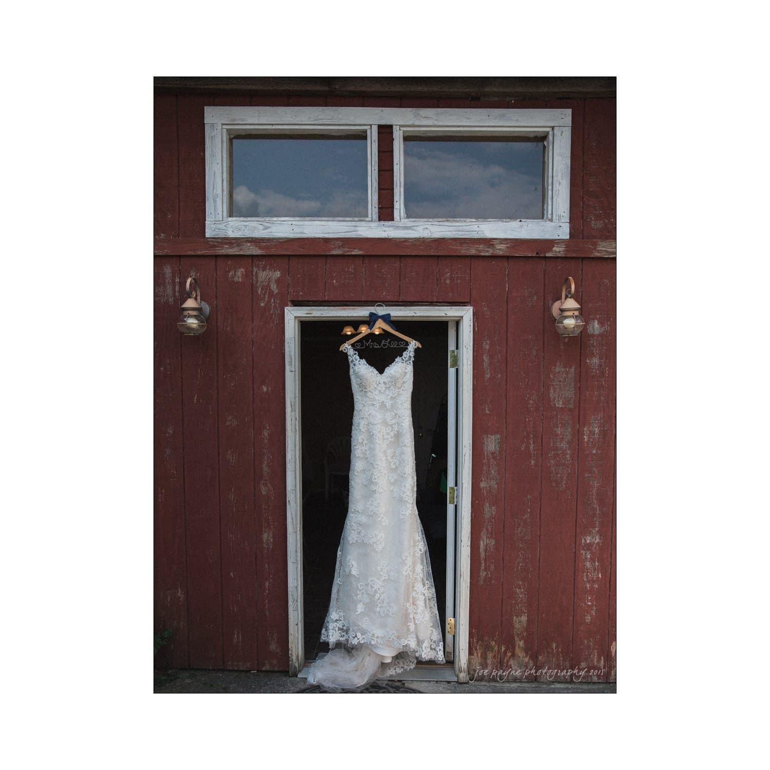 chapel hill wedding photography - nicole & wayne A lace wedding dress hangs on a hanger in the open doorway of a rustic red barn. Two lantern-style lights frame the entrance, and there are two rectangular windows above the door.