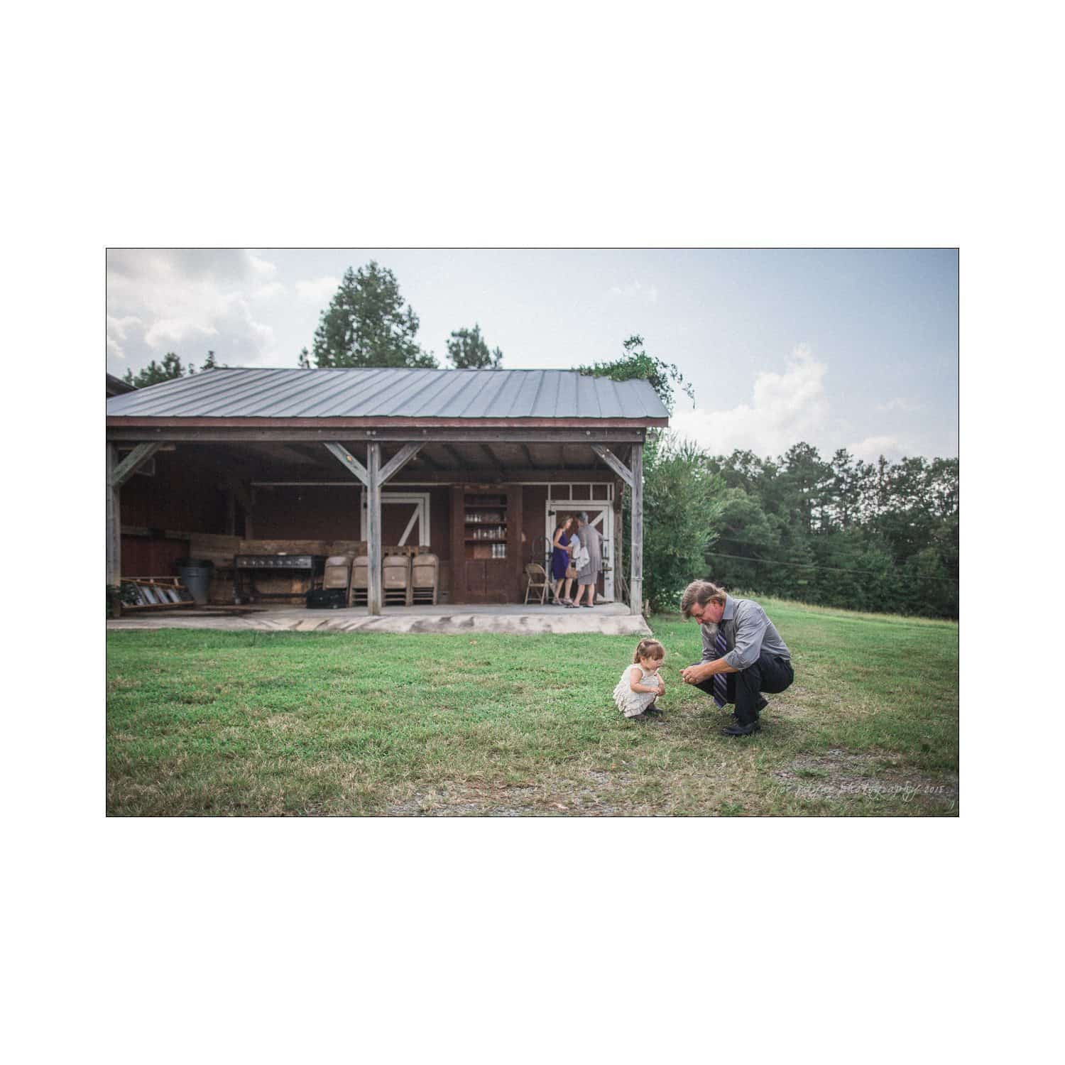 chapel hill wedding photography - nicole & wayne A man crouches on a grassy field, interacting with a small child. A wooden barn is in the background, and two people are visible near the entrance. The sky is partly cloudy, and the scene is serene and peaceful.