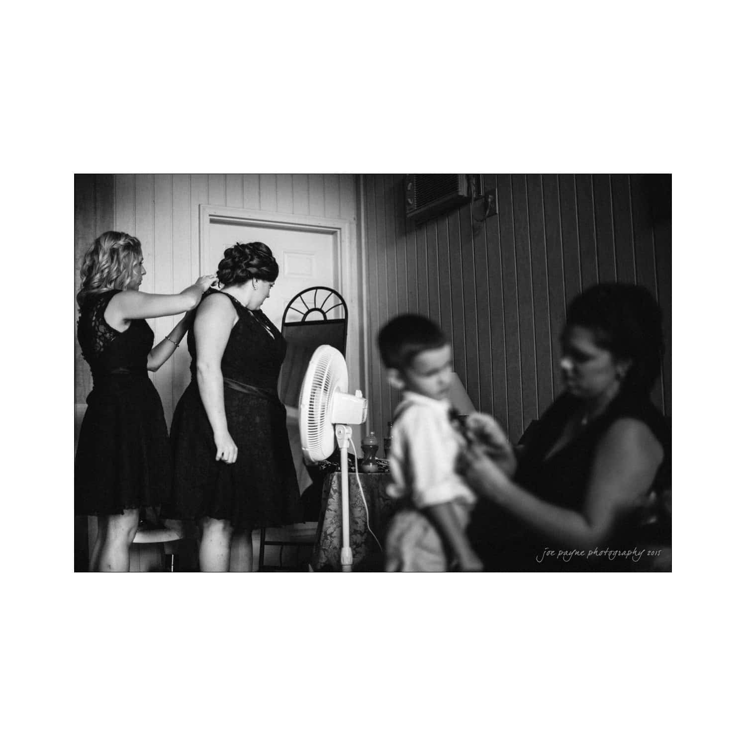 chapel hill wedding photography - nicole & wayne A black-and-white photo of a dressing room scene. Two women in elegant dresses are getting ready, with one helping adjust the others hair. Nearby, another woman is attending to a young boy. A fan and a door with a half-circle window are in the background.