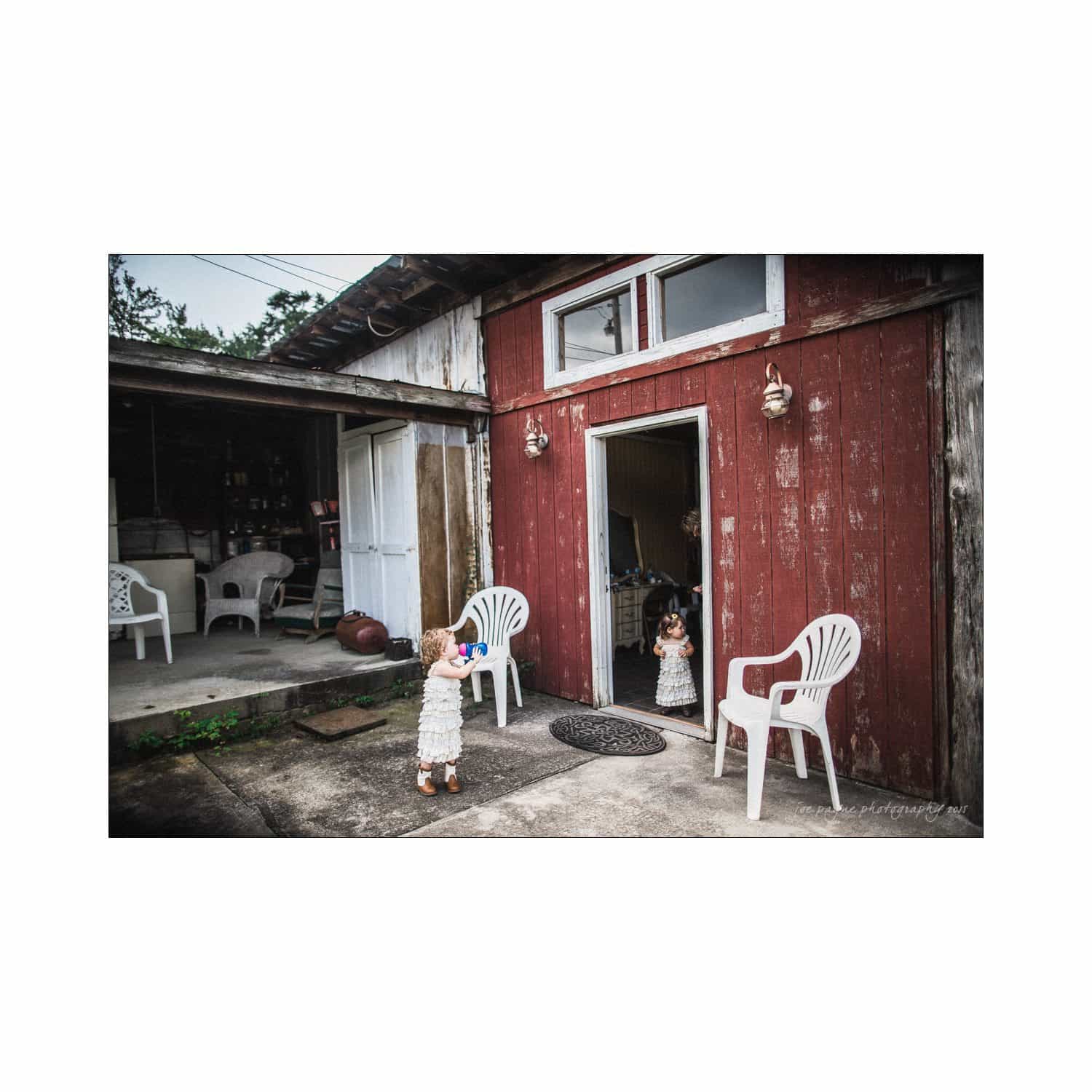 chapel hill wedding photography - nicole & wayne A child in a white dress stands outside a rustic red wooden building, looking at another child inside. Two white plastic chairs are placed nearby on the concrete. The scene has a rural and casual atmosphere.