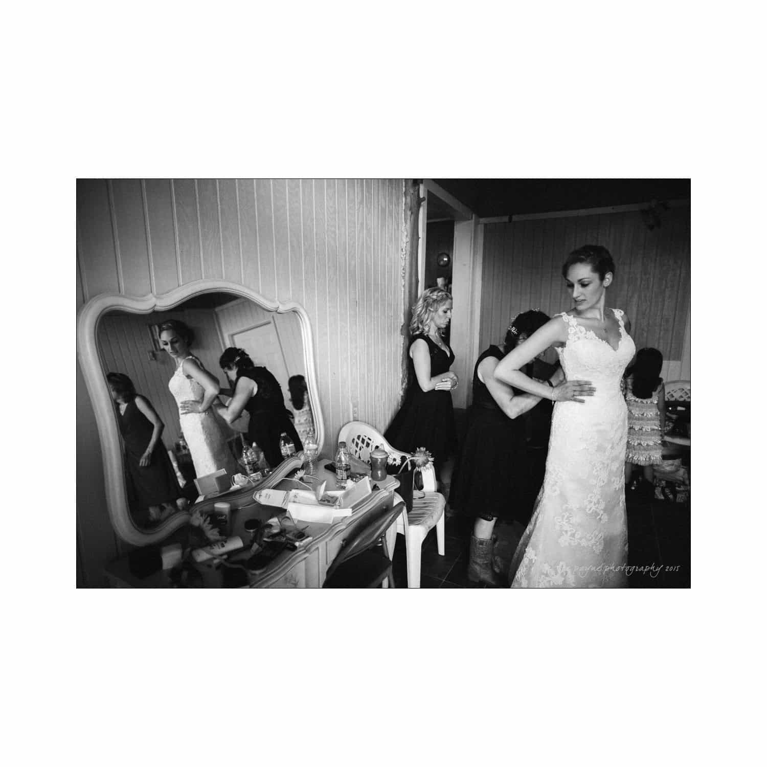 chapel hill wedding photography - nicole & wayne A bride in a lace gown stands confidently in front of a mirror while a woman assists her with adjustments. The room is cluttered with cosmetic and hair tools. Another woman is reflected in the mirror, wearing a dress and arranging her hair.