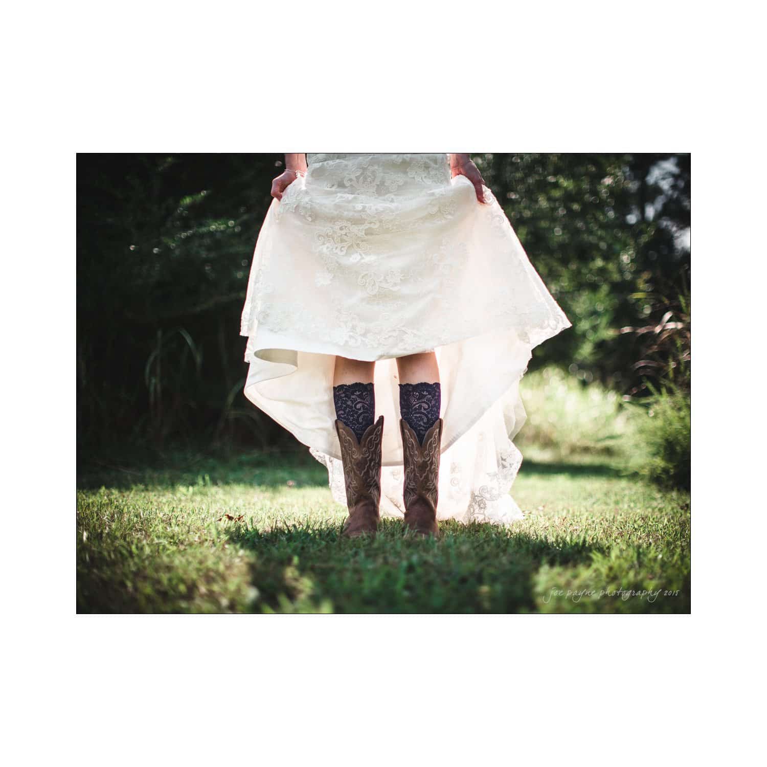 chapel hill wedding photography - nicole & wayne A bride stands on grass, wearing a white lace wedding dress lifted to reveal cowboy boots and lace stockings. The background is lush with greenery, suggesting a rustic outdoor setting.