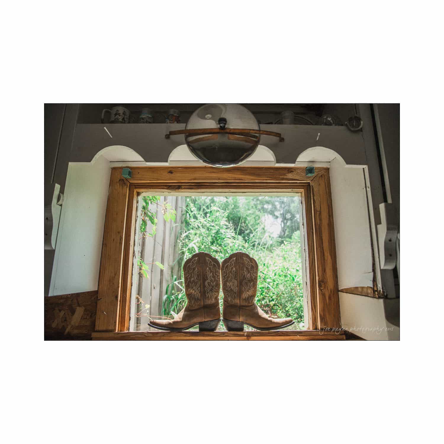 chapel hill wedding photography - nicole & wayne A pair of intricately detailed cowboy boots sit on a windowsill. The window is open with rustic wooden shutters, framing a lush green outdoor view. Soft natural light illuminates the boots and the interior around them.