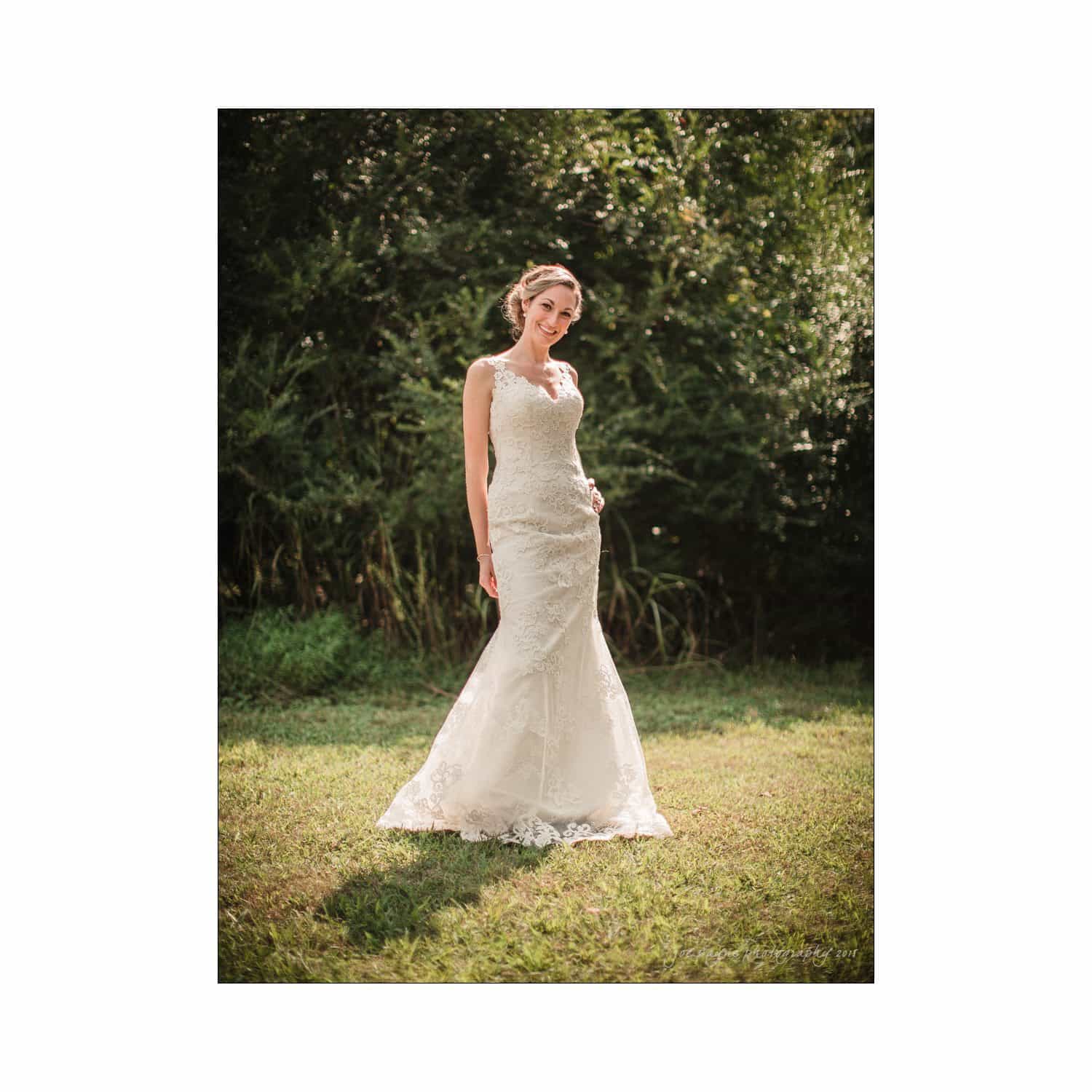 chapel hill wedding photography - nicole & wayne A person wearing a fitted lace wedding gown stands outdoors on a grassy area, surrounded by lush green foliage. The gown features a sleeveless design with intricate detailing. The background is softly blurred, emphasizing the serene natural setting.