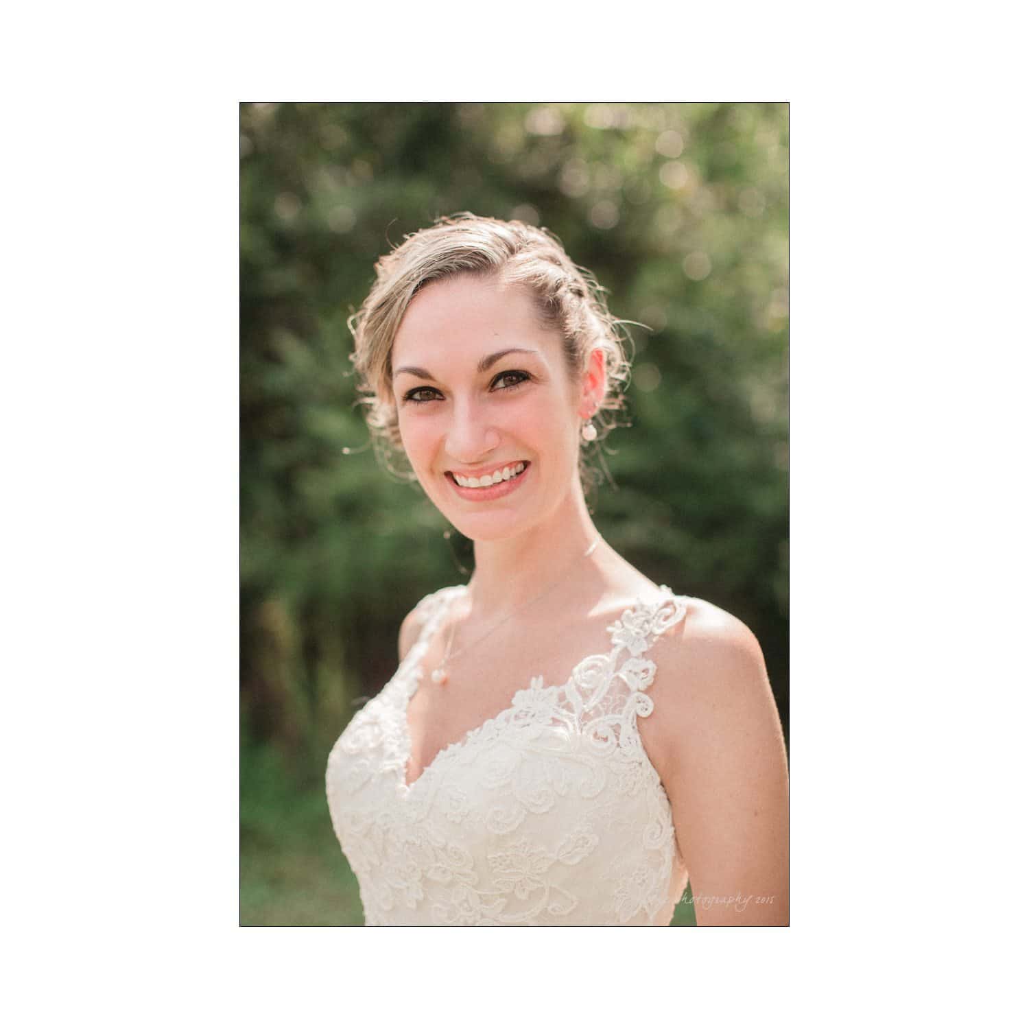 chapel hill wedding photography - nicole & wayne A smiling woman in a lace wedding dress stands outdoors with sunlight illuminating her. She has short, wavy hair styled with an elegant hairpiece and is wearing subtle jewelry. The background is blurred greenery.