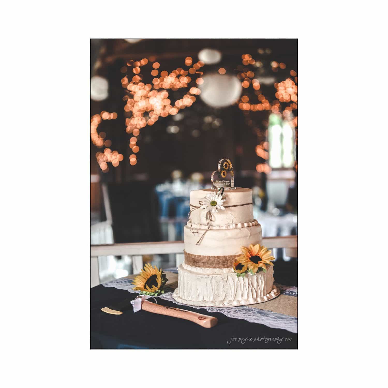 chapel hill wedding photography - nicole & wayne A three-tiered white wedding cake decorated with burlap ribbon and sunflowers, topped with a number 60 ornament. The cake is centered on a table with blurred bokeh lights and a dimly lit reception hall in the background.