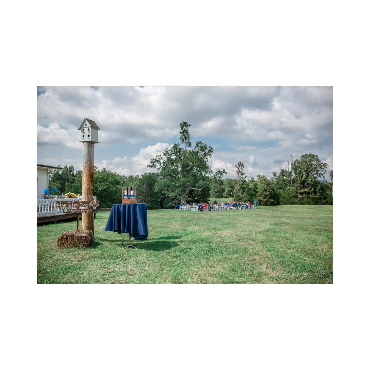 chapel hill wedding photography - nicole & wayne A grassy field with a birdhouse on a tall pole to the left and a small podium covered with a blue cloth. In the background, a group of people is gathered, seated on white chairs under a partly cloudy sky.