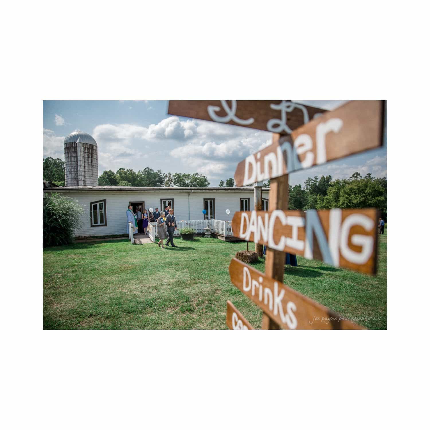 chapel hill wedding photography - nicole & wayne A rustic outdoor scene with a wooden sign pointing to dinner, dancing, and drinks. A group of people stands near a barn-like building with a silo in the background, set against a partly cloudy sky.