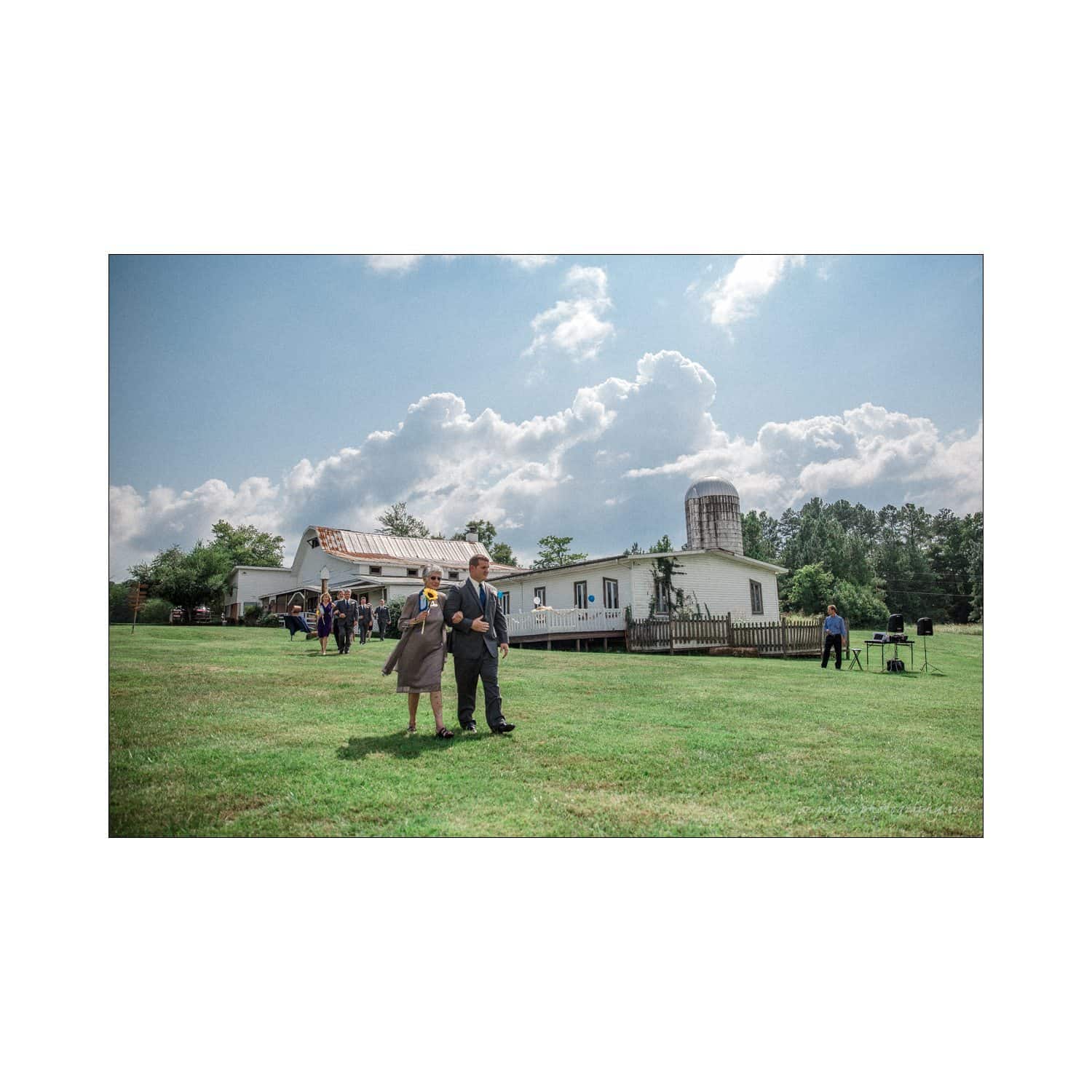 chapel hill wedding photography - nicole & wayne A couple stands on a grassy field with a rustic white barn and silo in the background. Puffy clouds dot the blue sky, while a few people and a camera setup are visible at a distance.