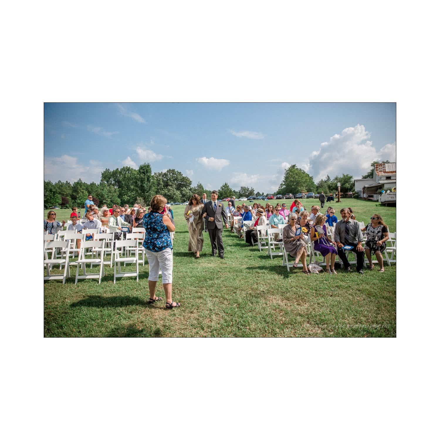 chapel hill wedding photography - nicole & wayne A woman stands and addresses a seated crowd outdoors. People are sitting and listening, with trees and a house in the background under a partly cloudy sky.