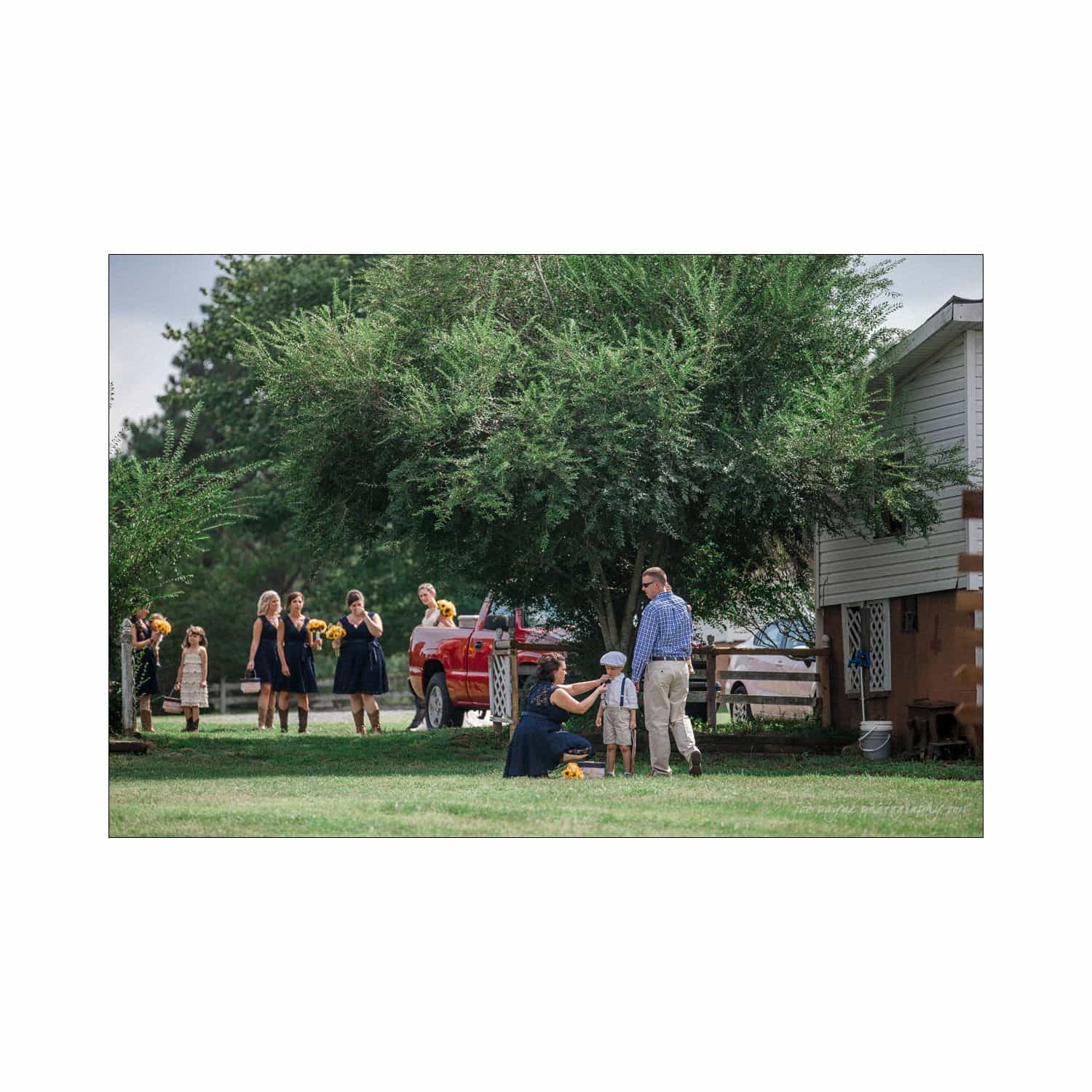 chapel hill wedding photography - nicole & wayne A group of people in casual attire are gathered outdoors on a grassy area beside a house and trees. A pickup truck is parked nearby. The scene appears relaxed and social.