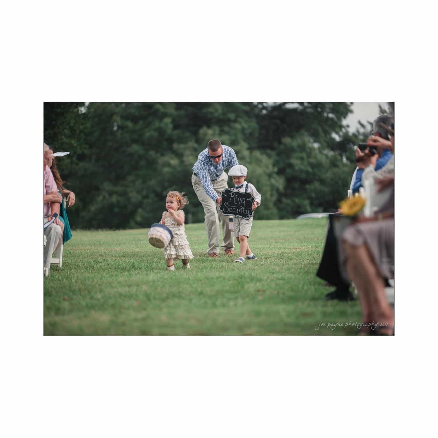 chapel hill wedding photography - nicole & wayne Two young children walk down an outdoor aisle during a wedding. One child is carrying a sign that says Ring Security, and the other holds a basket. An adult behind them is smiling as guests seated on either side watch.