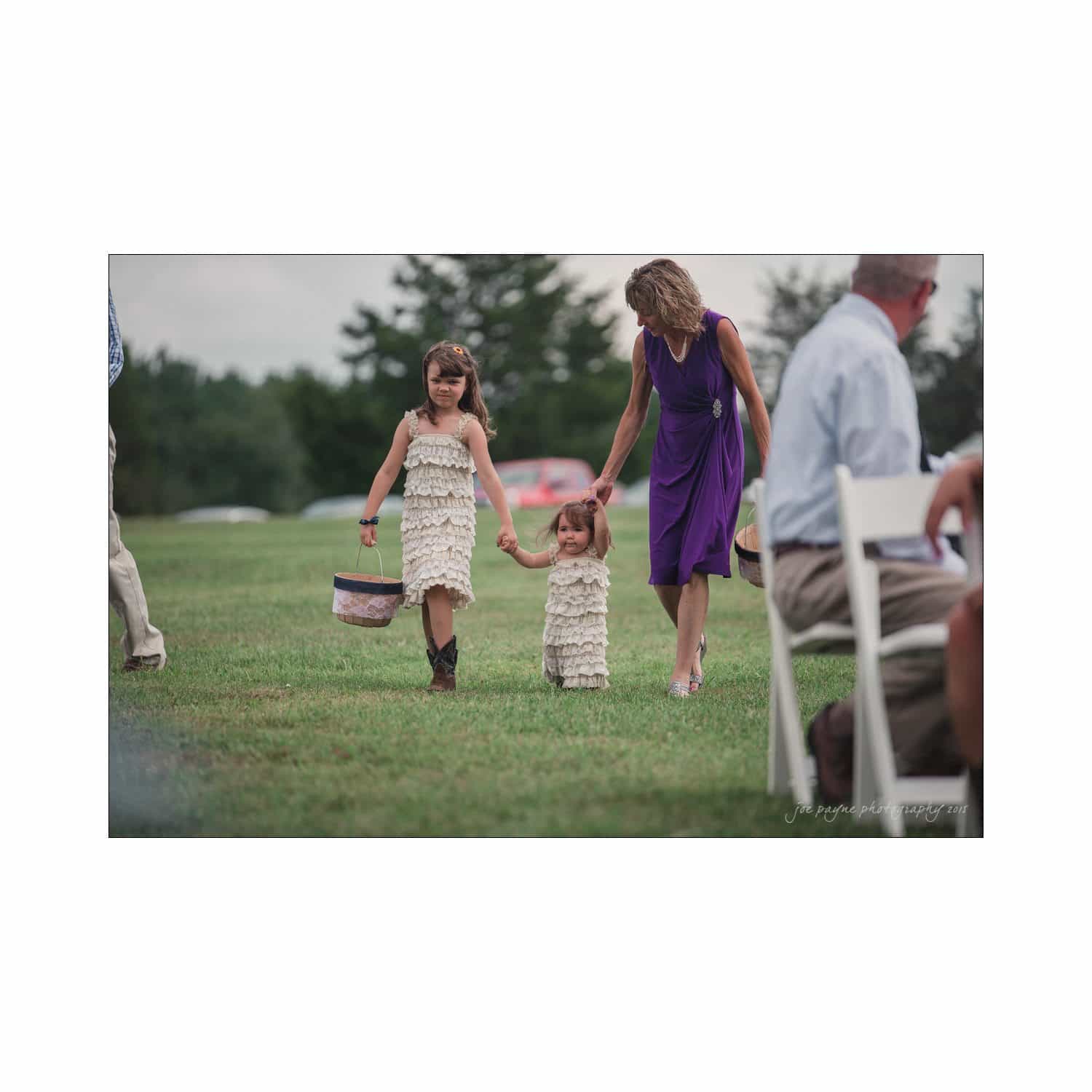 chapel hill wedding photography - nicole & wayne A woman in a purple dress walks across a grassy area with two young girls. The girls are wearing matching ruffled dresses and holding hands, with one carrying a basket. People are seated nearby.