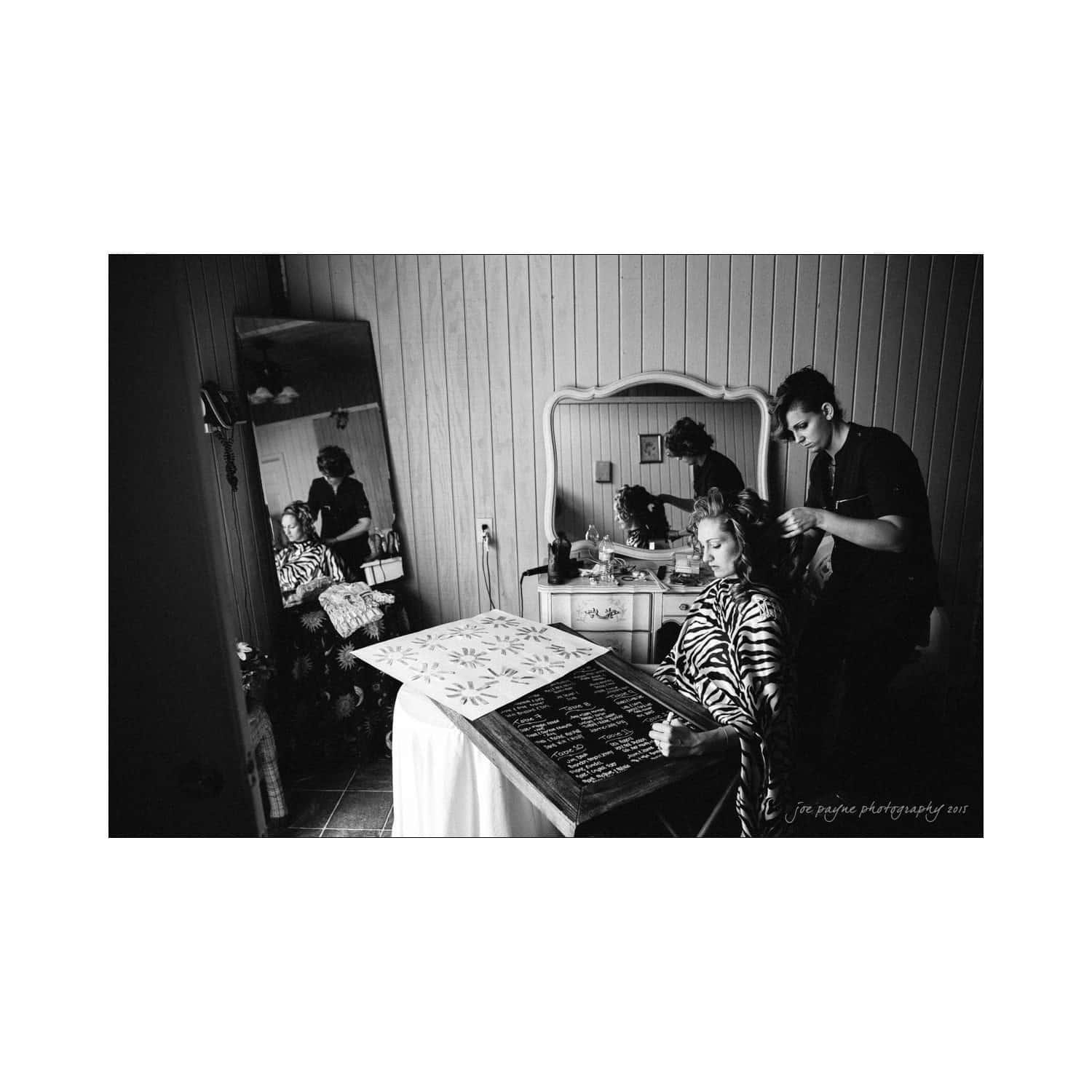 chapel hill wedding photography - nicole & wayne Black and white photo of a woman having her hair styled by a hairdresser in a room with wooden walls. She sits in front of a mirror, and theres a decorated surface with writing on it nearby.