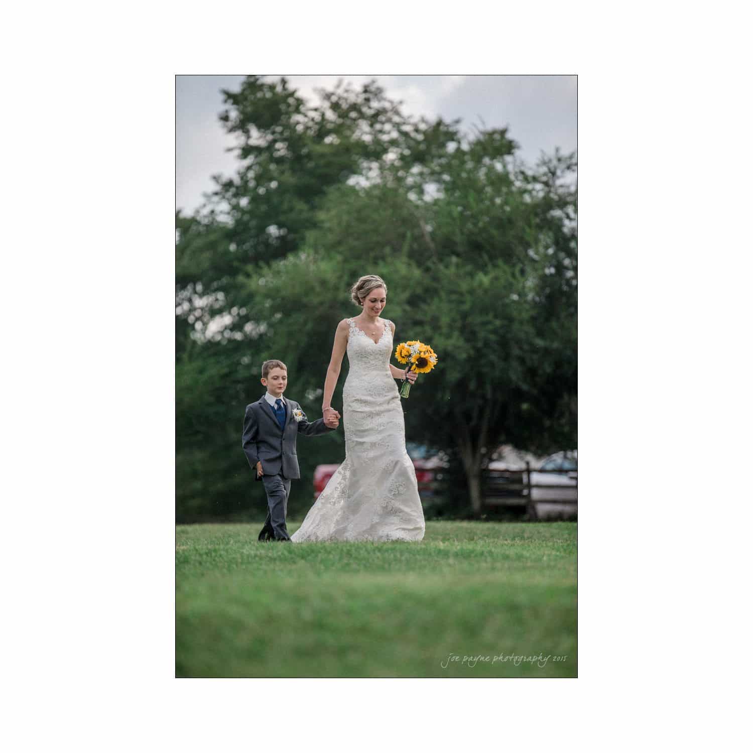 chapel hill wedding photography - nicole & wayne A woman in a white wedding dress holds a bouquet of sunflowers and walks hand in hand with a young boy in a suit across a grassy field, with trees in the background.