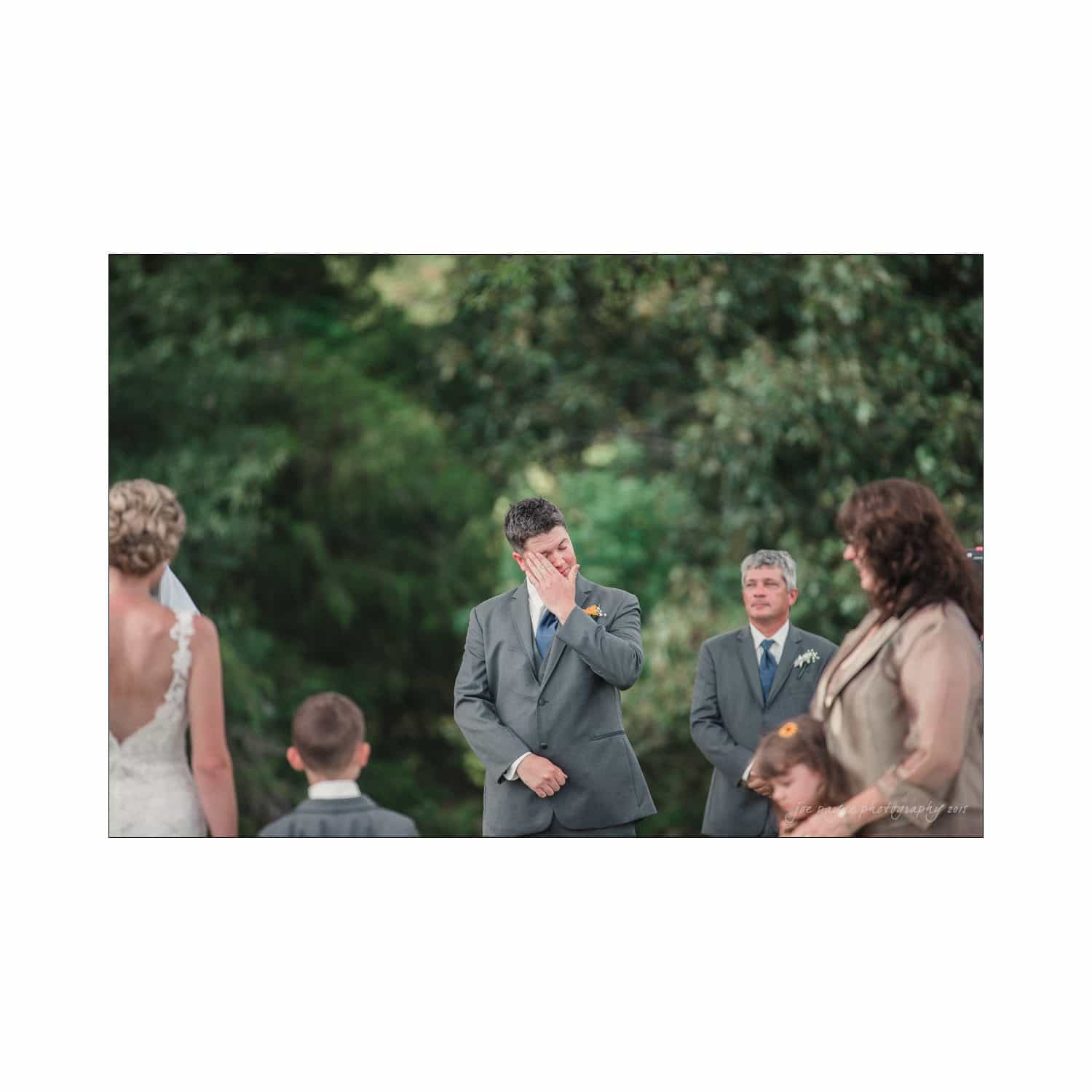 chapel hill wedding photography - nicole & wayne A groom in a gray suit wipes tears during an outdoor wedding ceremony. The bride, dressed in a white gown, stands across from him. A boy, a man in a suit, and a woman holding a child are present. The background is lush greenery.