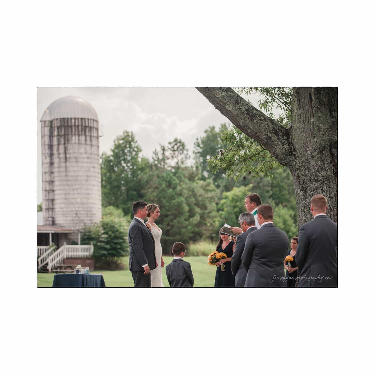 chapel hill wedding photography - nicole & wayne A wedding ceremony outdoors near a large tree and an old silo. The bride and groom stand facing each other, surrounded by their wedding party. A young boy holds a bouquet. The atmosphere is serene and intimate, with a lush green landscape in the background.