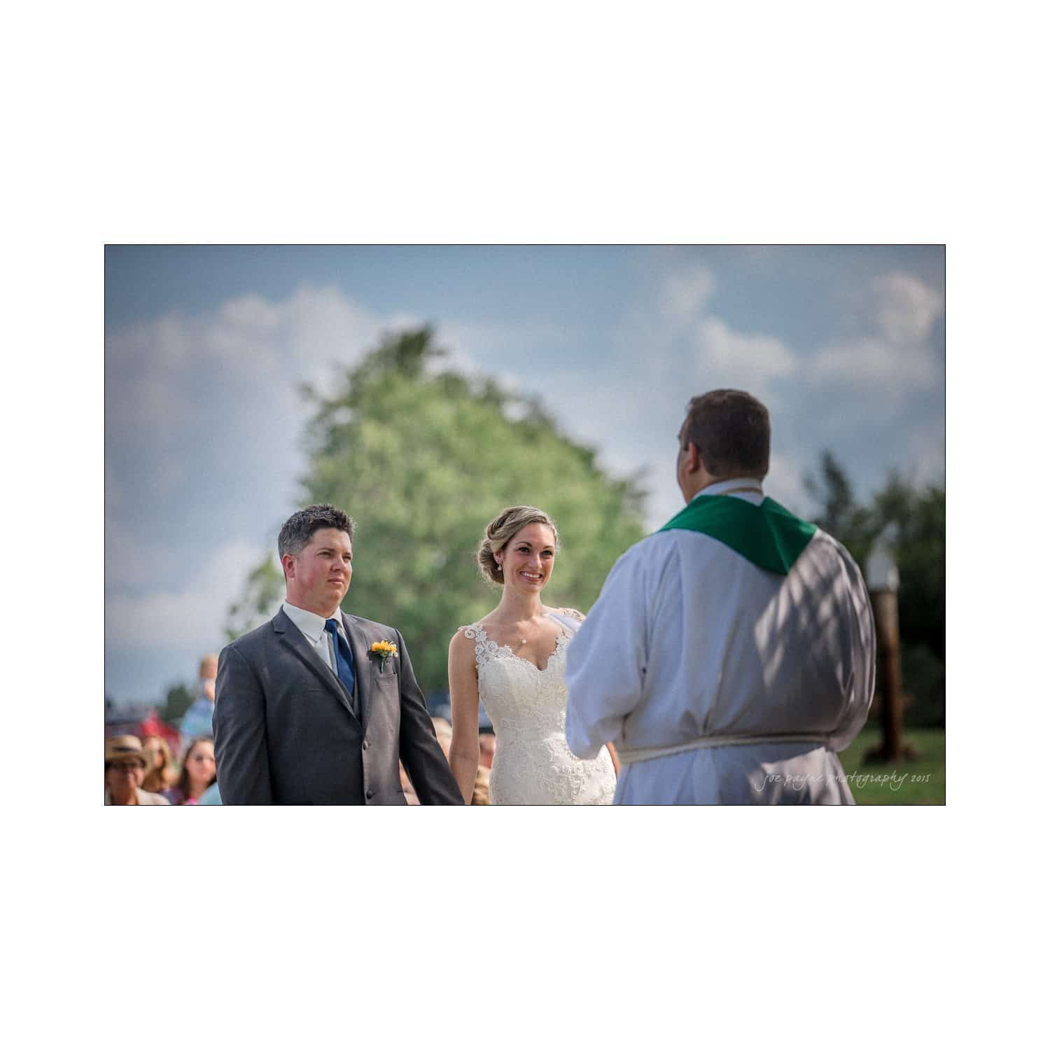 chapel hill wedding photography - nicole & wayne A bride and groom stand facing a clergyman during an outdoor wedding ceremony. The bride wears a lacy white gown, and the groom is in a gray suit with a flower boutonniere. Guests are seated in the background under a clear blue sky.