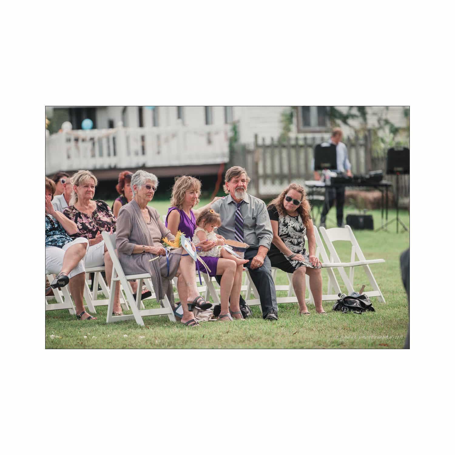 chapel hill wedding photography - nicole & wayne A group of people sit on white folding chairs during an outdoor event. The scene includes adults and a child, all casually dressed. In the background, a musician stands near audio equipment. The setting is grassy with a white building nearby.
