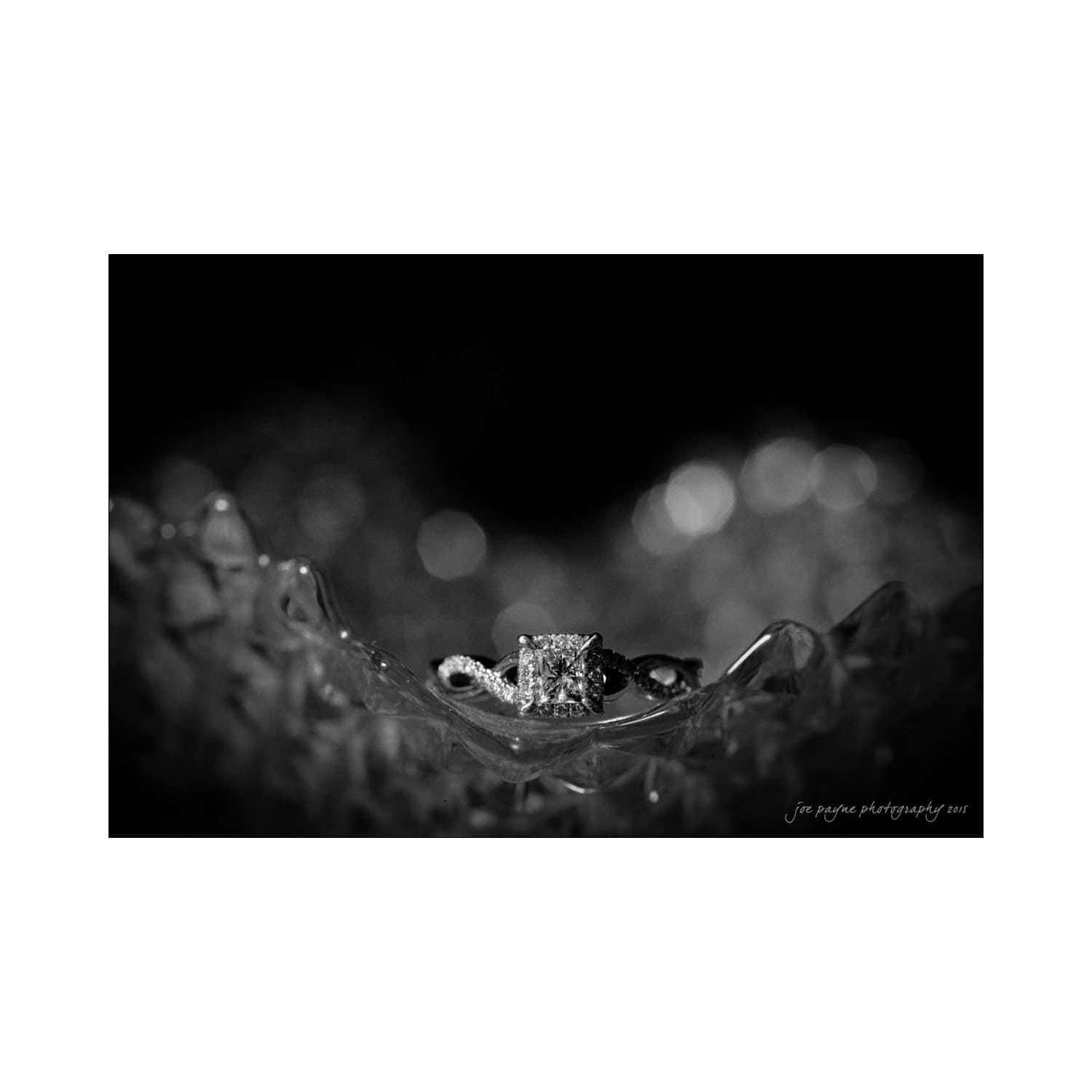 chapel hill wedding photography - nicole & wayne A black and white photo of an engagement ring with a square diamond, elegantly placed on a textured, sparkling surface. The background is softly blurred to highlight the rings intricate details.
