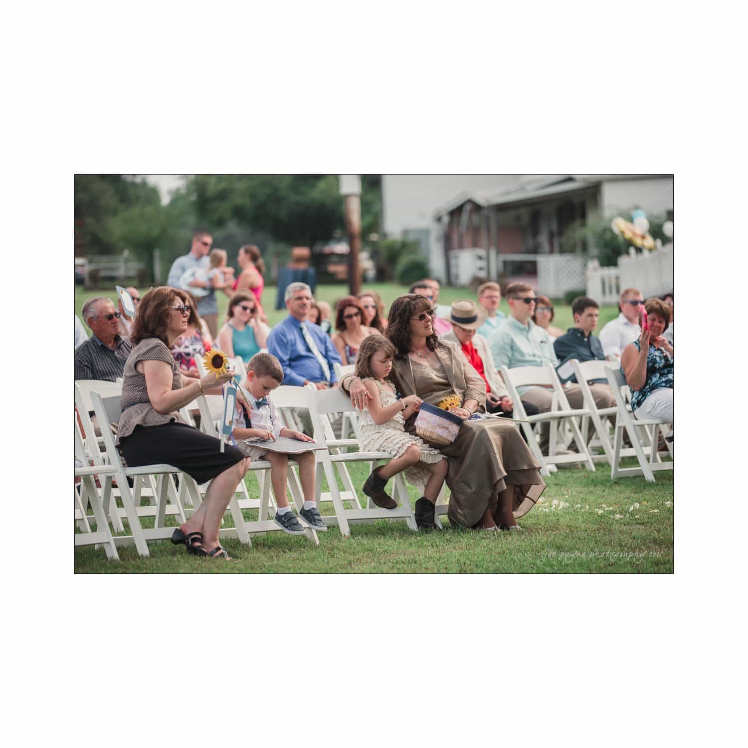 chapel hill wedding photography - nicole & wayne People seated outdoors on white folding chairs attending a gathering. A woman with sunflowers sits with two children. Others chat and observe the event. The background shows trees and a building. It’s a sunny day.