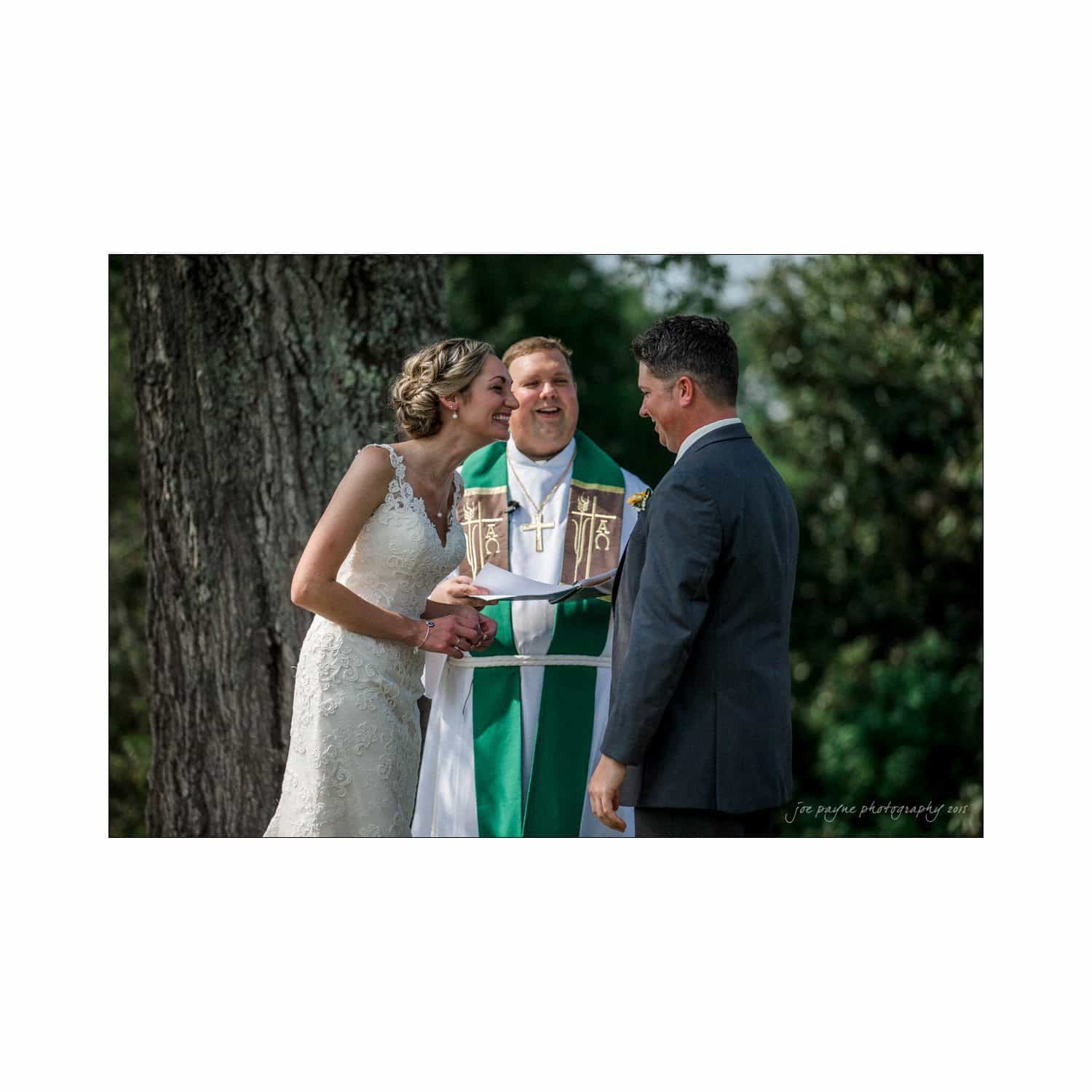 chapel hill wedding photography - nicole & wayne A bride and groom stand beside a smiling officiant wearing a green and white stole during an outdoor wedding ceremony. The bride leans towards the officiant, laughing. A tree and blurred greenery are in the background.