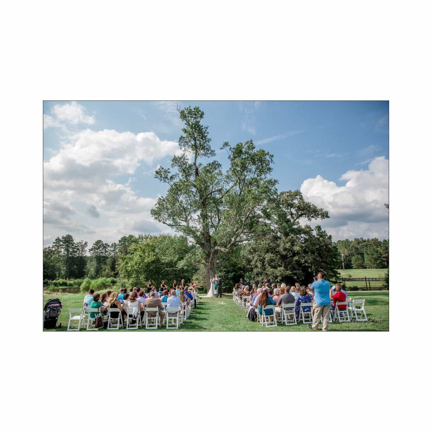 chapel hill wedding photography - nicole & wayne A wedding ceremony is taking place outdoors under a large tree. Guests are seated in white chairs on either side of the aisle. The setting is a lush, green landscape with a clear blue sky and scattered clouds.
