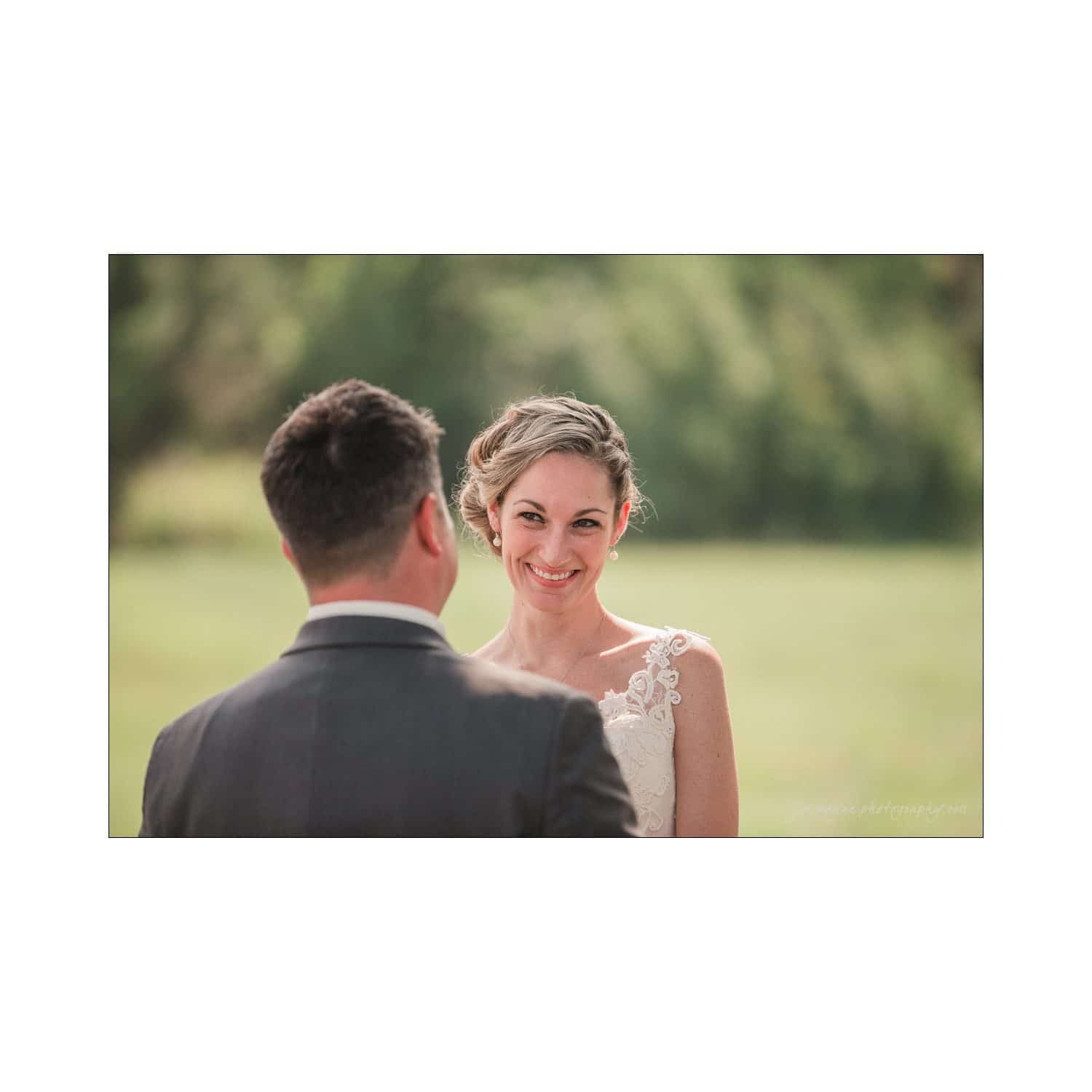 chapel hill wedding photography - nicole & wayne A smiling bride with a lace dress and braided hairstyle looks at a groom in a suit during an outdoor wedding ceremony. The background is a blurred green landscape.