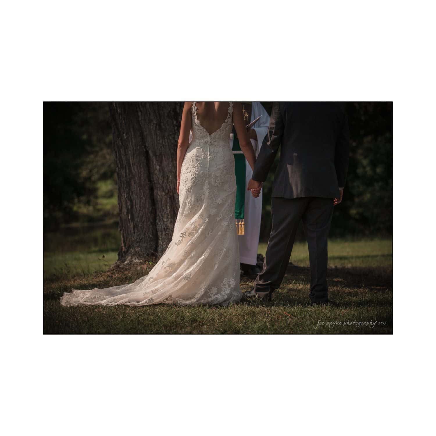 chapel hill wedding photography - nicole & wayne A bride and groom stand side by side outdoors in front of a tree. The bride wears a long, lace wedding dress, and the groom is in a dark suit. A person in white attire stands behind them, possibly officiating the ceremony.