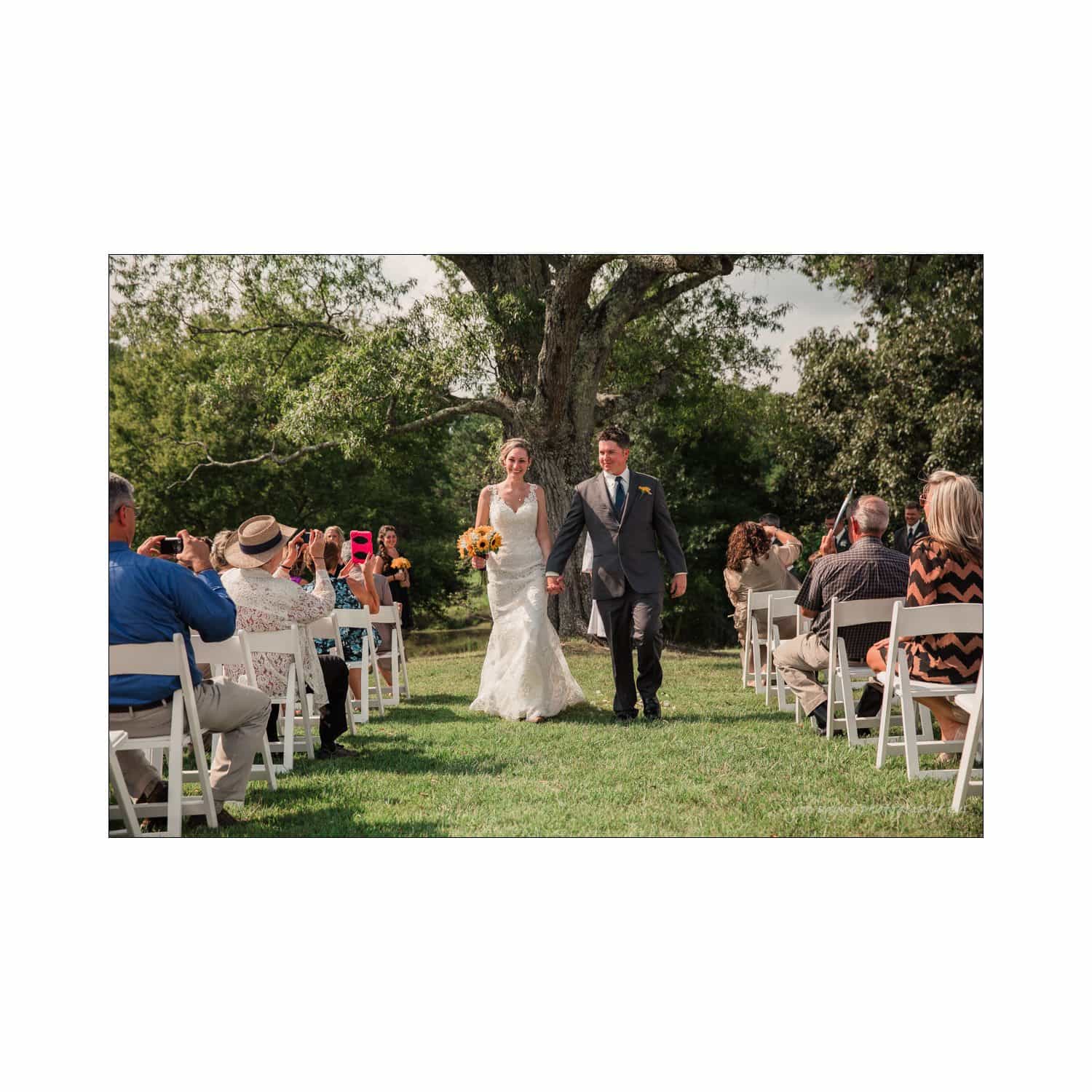 chapel hill wedding photography - nicole & wayne A bride and groom walk down an outdoor aisle, smiling, as guests seated on white chairs applaud. The bride holds a bouquet of yellow flowers, and the groom wears a dark suit. Greenery and trees can be seen in the background under a sunny sky.