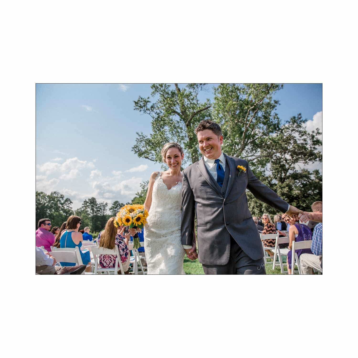 chapel hill wedding photography - nicole & wayne A smiling couple, dressed in a wedding gown and suit, walk hand in hand down an outdoor aisle. The bride holds a bouquet of sunflowers as guests seated in white chairs look on, with a backdrop of trees and a blue sky.