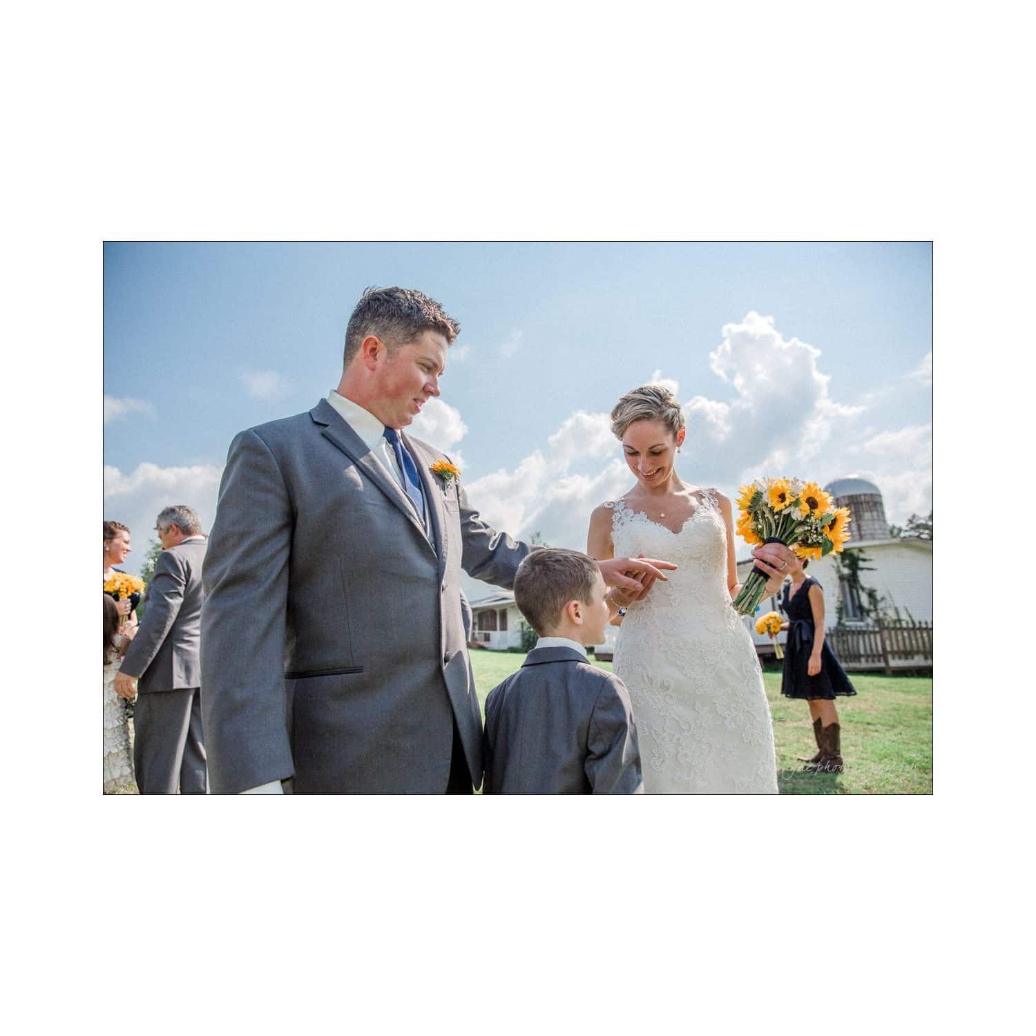 chapel hill wedding photography - nicole & wayne A bride and groom stand outside on a sunny day, smiling at a child in front of them. The bride holds a bouquet of sunflowers, while other people in formal attire are visible in the background against a blue sky with clouds.