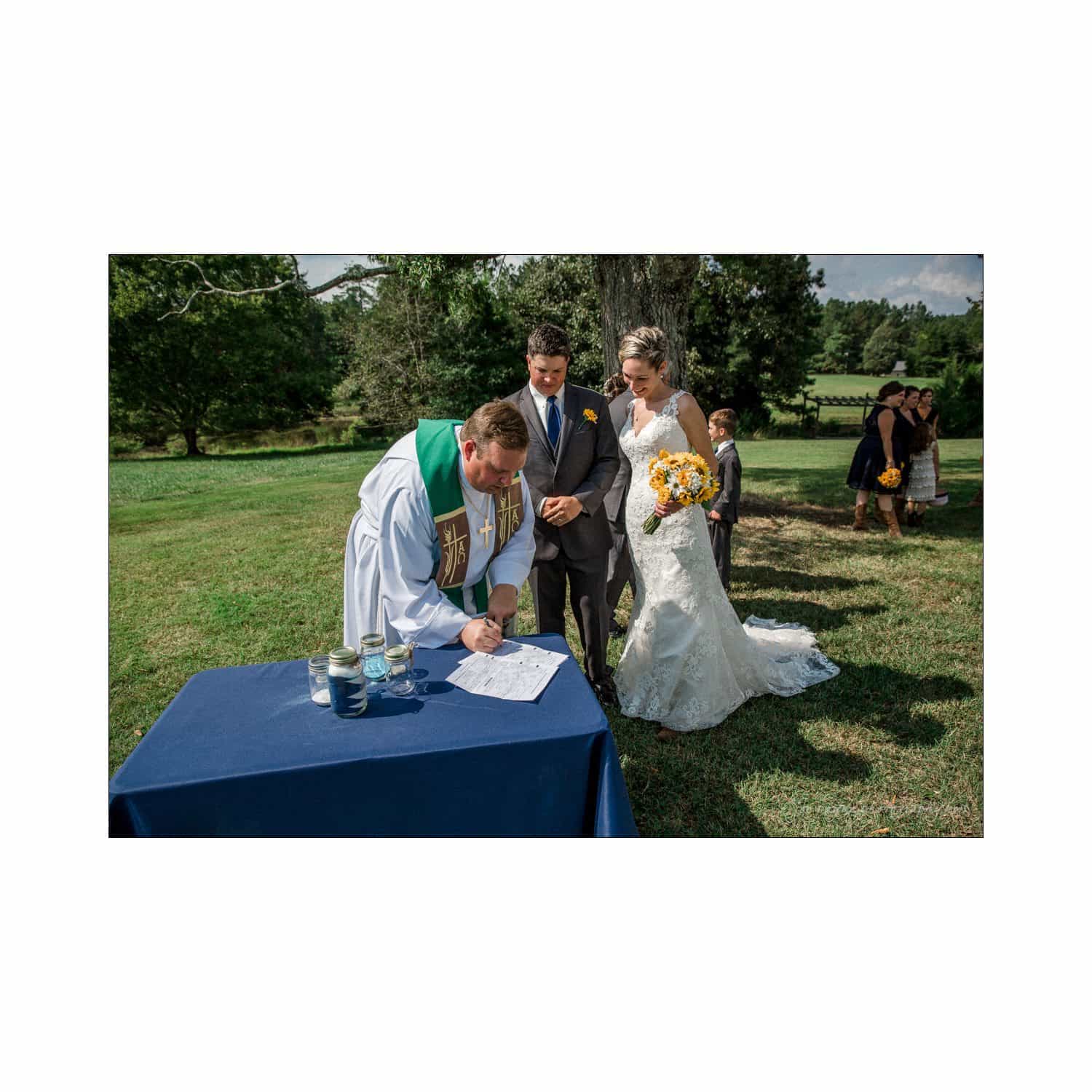 chapel hill wedding photography - nicole & wayne A bride and groom stand outdoors near a table. The groom holds a bouquet while a person in a religious robe signs a document. Trees and guests are in the background on a sunny day.