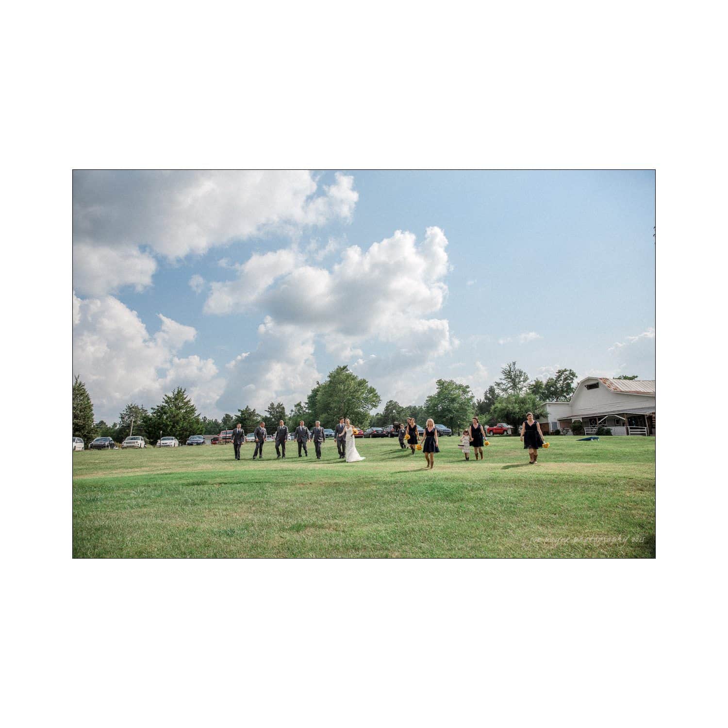 chapel hill wedding photography - nicole & wayne A bridal party walks in a green field under a partly cloudy sky. The group is dressed in formal attire, with a bride in white at the center. Cars and trees are visible in the background near a large building.