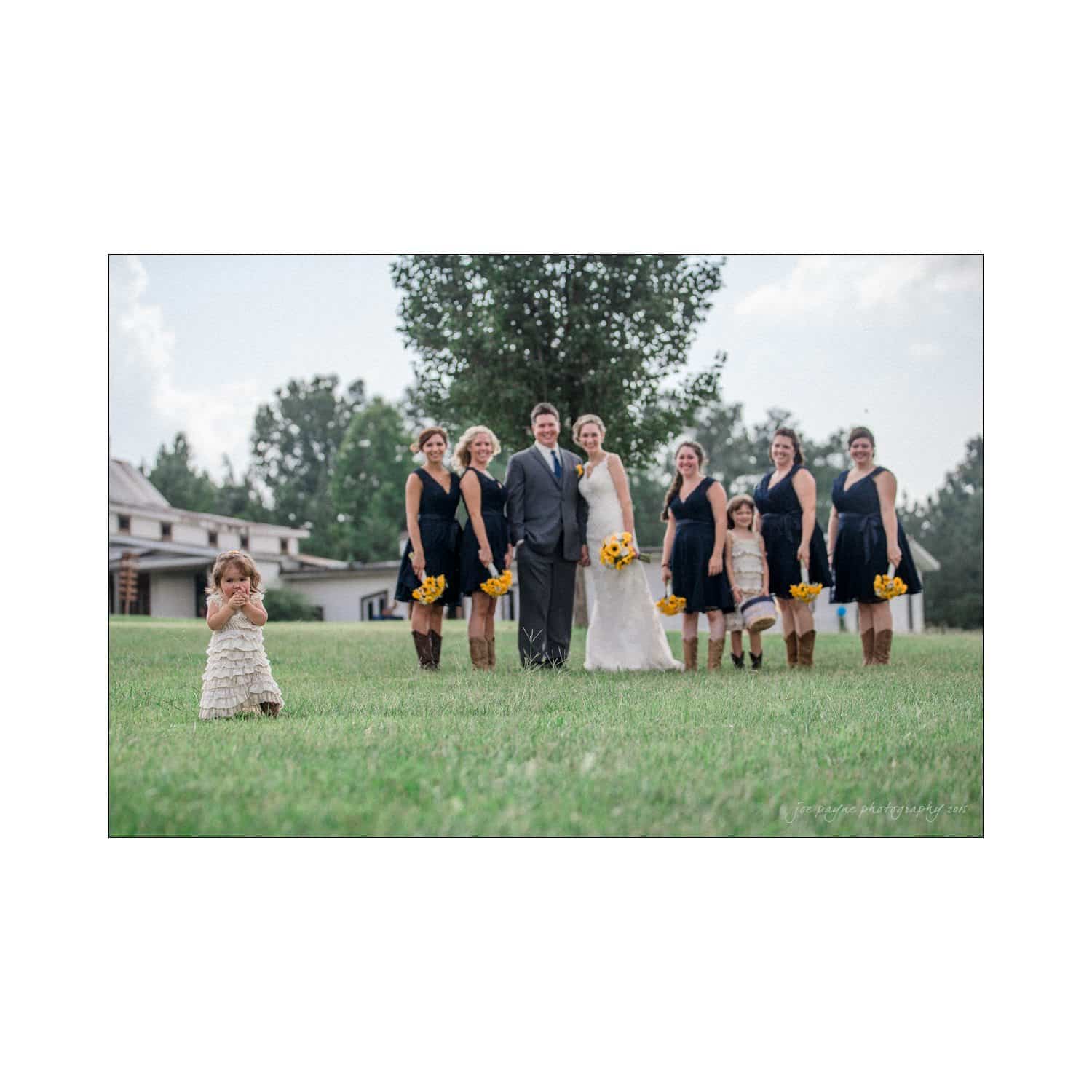 chapel hill wedding photography - nicole & wayne A bride and groom stand with their wedding party on a grassy field. The bridesmaids wear black dresses and hold yellow bouquets. A child in a white dress stands to the side. Trees and a building are in the background.