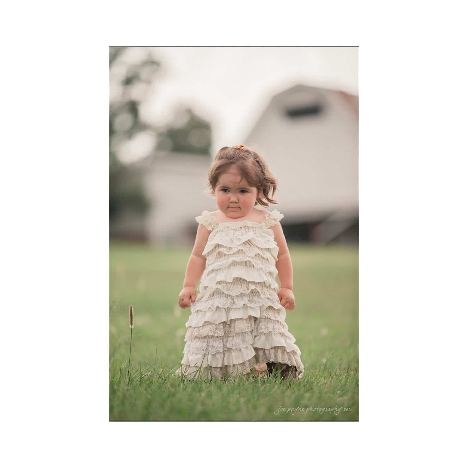 chapel hill wedding photography - nicole & wayne A toddler in a frilly white dress stands on grass in front of a blurred background that includes a barn. The child looks directly at the camera with a neutral expression.