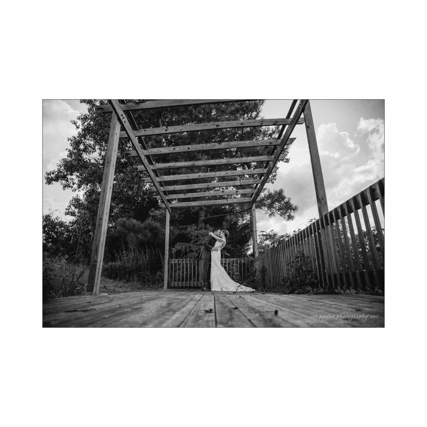 chapel hill wedding photography - nicole & wayne A black and white photo of a couple embracing under a wooden pergola on a deck. The bride wears a long gown, and the groom is in a suit. The background includes trees and a cloudy sky.