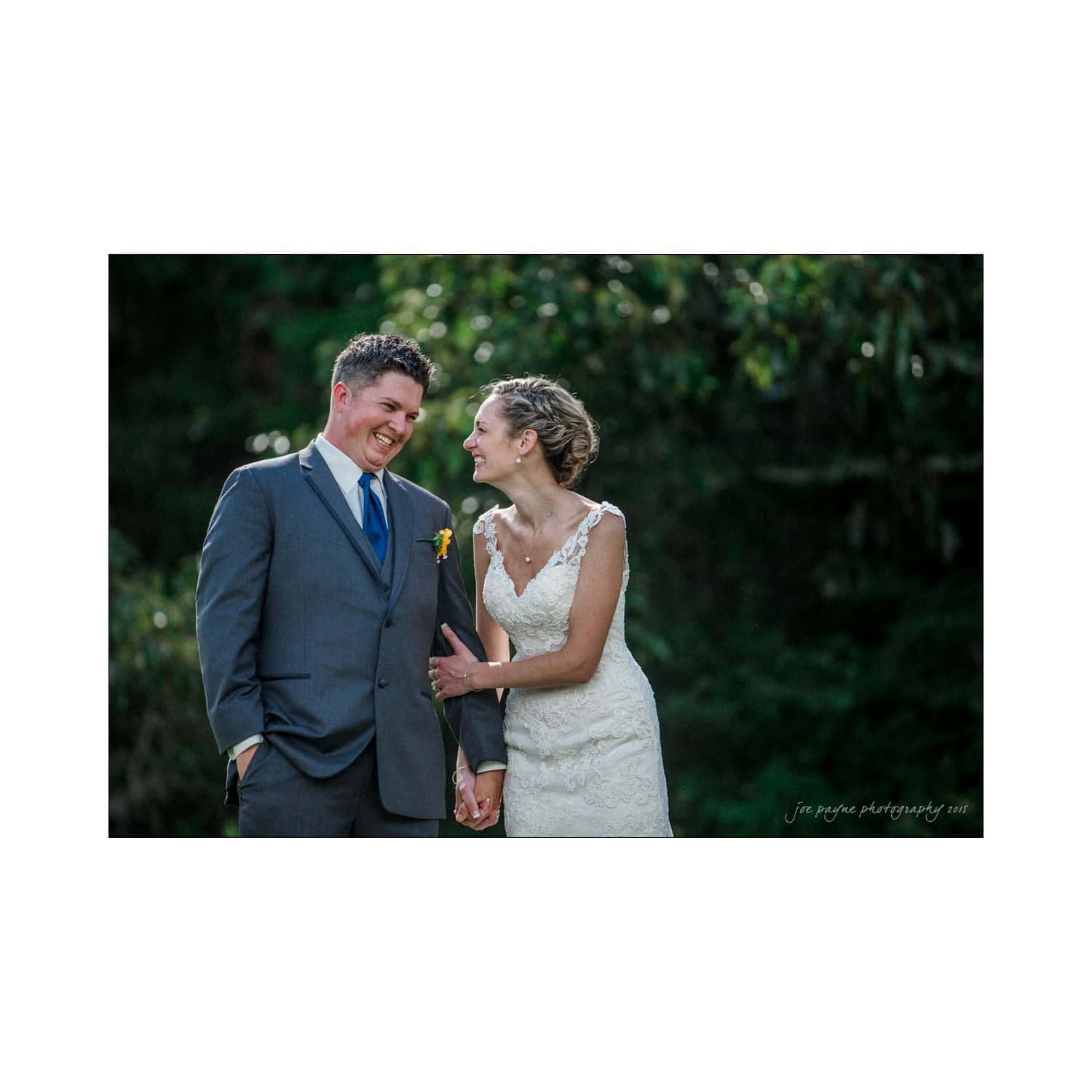 chapel hill wedding photography - nicole & wayne A bride in a white lace dress and a groom in a gray suit share a joyful moment, holding hands and laughing outdoors, with greenery in the background.
