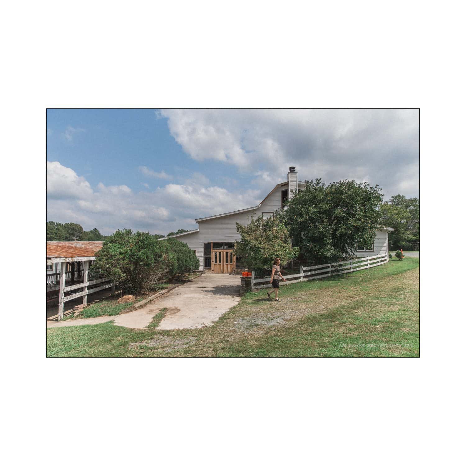 chapel hill wedding photography - nicole & wayne A person walks along a pathway in front of a white building with a sloped roof, surrounded by trees and grass. Its a sunny day with scattered clouds in the sky.